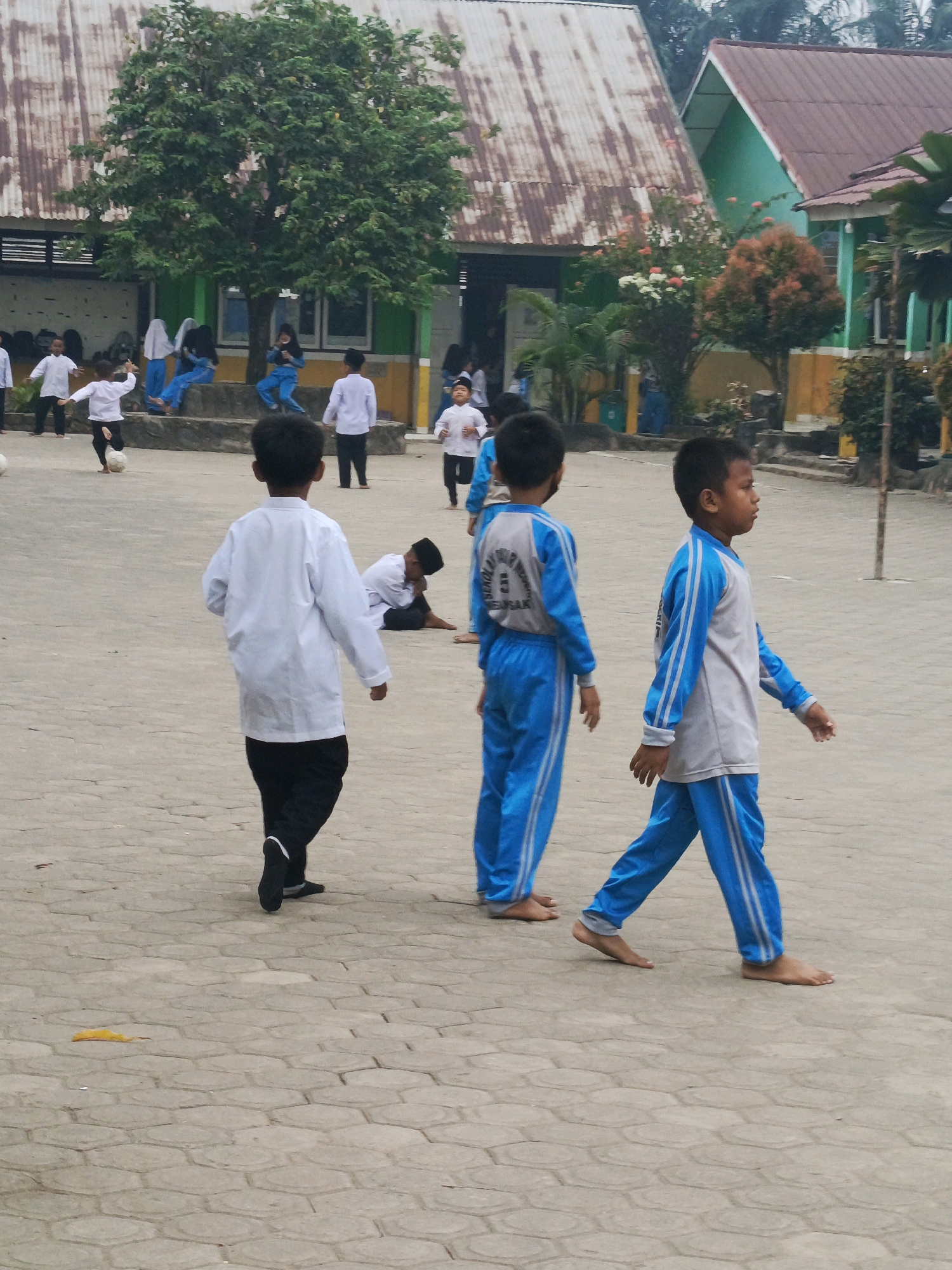 The image depicts a group of young people, likely students, in an outdoor courtyard area. In the foreground, three boys are walking. The boy on the left is wearing a white long-sleeved shirt and black pants. The middle boy is wearing a gray long-sleeved shirt with blue sleeves and blue pants. The boy on the right is also wearing a gray long-sleeved shirt with blue sleeves and blue pants, and he appears to be looking to his right. Behind them, another boy in a white shirt and black pants is partially visible, and a boy in a white shirt and black pants is sitting on the ground with his head bowed. Other individuals, including those wearing hijabs, are visible in the background, some sitting and some standing. A soccer ball is visible on the ground near the left side of the image. The environment includes a paved courtyard with a hexagonal pattern, buildings with corrugated metal and tiled roofs in the background, and several trees, one large and leafy. The overall scene suggests a school or community gathering in a tropical or subtropical climate, consistent with a location like Megang Sakti II, Indonesia.