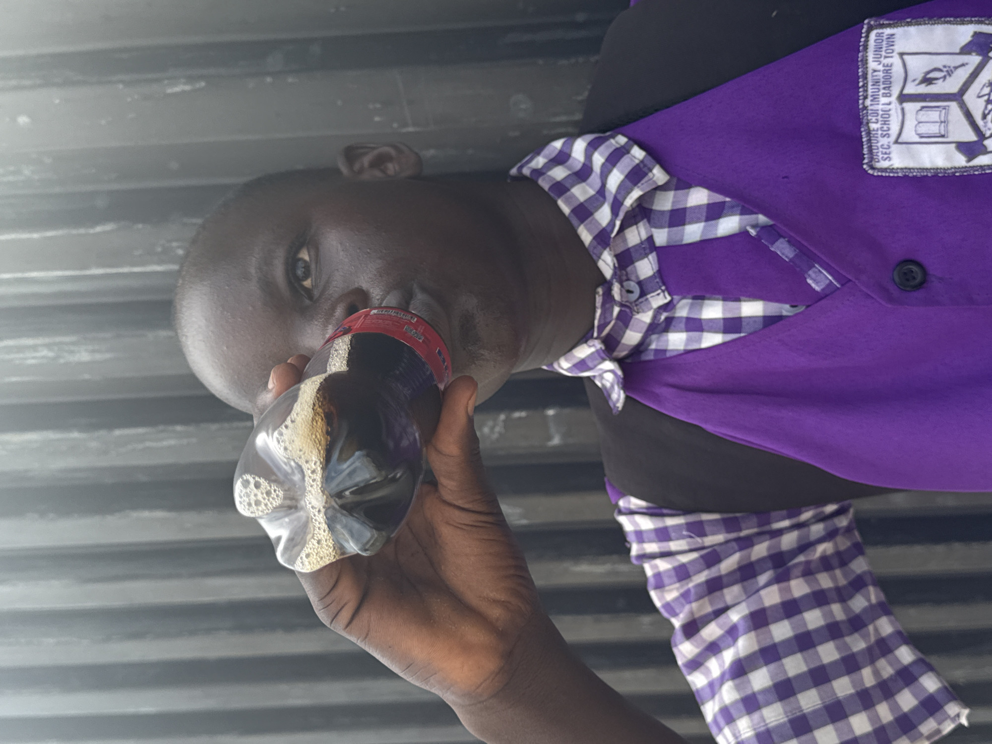 A man drinks from a plastic bottle outdoors in Ado, Nigeria. The dark-skinned man is wearing a purple collared shirt with white and purple checkered sleeves and a patch on the upper right that reads "Badore Community Junior Sec. School Badore Town." He is holding a plastic bottle filled with a dark liquid in his right hand, tilting the bottle to drink. The background is a corrugated metal wall. The image is rotated counterclockwise.