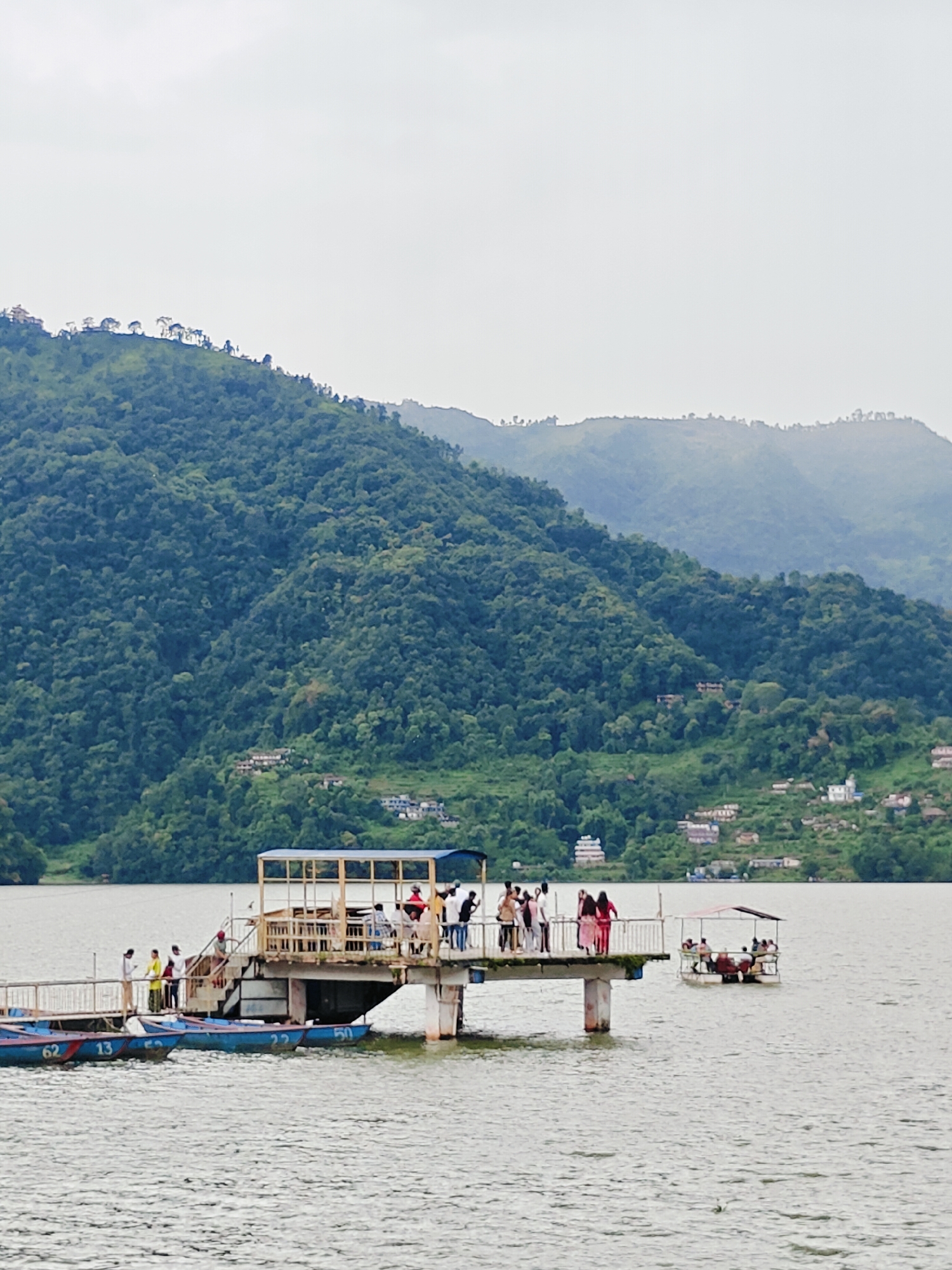 The image captures a serene lakeside scene in Pokhara, Nepal, likely at Phewa Lake, given the prominent view of lush, tree-covered hills characteristic of the region. In the foreground, several blue boats are moored to a pier, some displaying numbers like "62", "13", "22", and "50". A group of people are standing on the pier, some interacting with each other, while others gaze out at the water. A second, smaller boat with a canopy and several passengers is visible further out on the lake. The background is dominated by a majestic, verdant mountain range, its slopes dotted with scattered houses, suggesting a settled, yet wild, landscape. The sky is overcast, contributing to a soft, diffused light that bathes the entire scene.