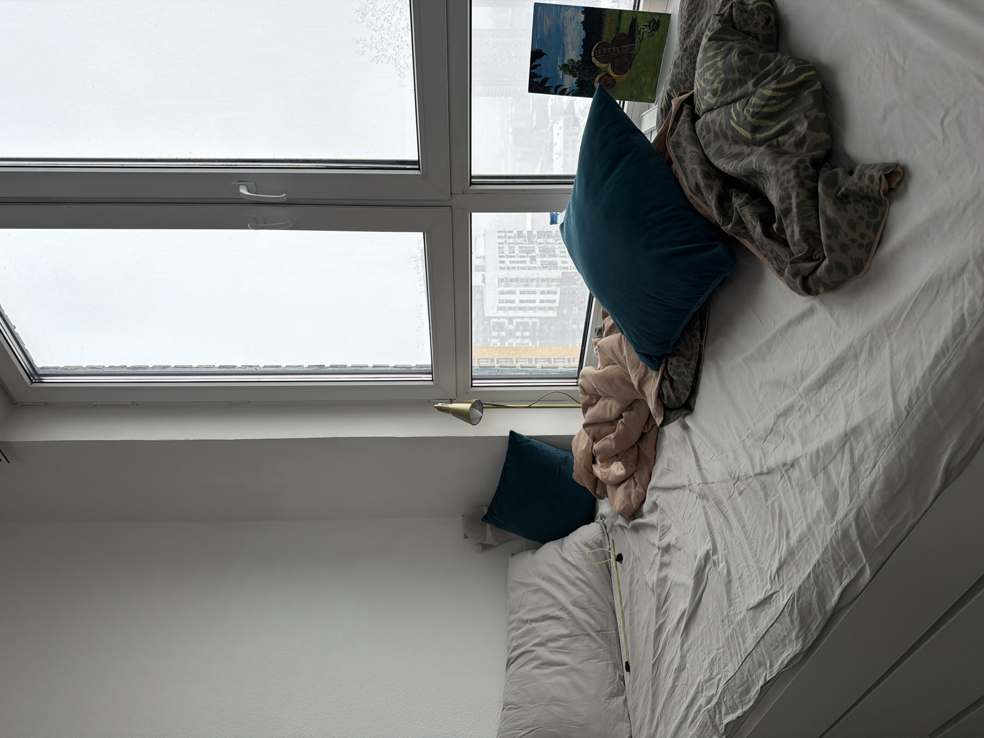 An overhead interior shot depicts an unmade bed positioned directly beneath a large window. The bed is covered with light grey sheets, showing numerous wrinkles and folds. Two dark teal-colored pillows are placed at the head of the bed, one larger and one smaller, alongside a light grey pillow. A crumpled, beige-pink duvet or blanket lies on the bed, partially covered by another blanket featuring a muted grey and green animal print pattern, likely leopard or cheetah.
The multi-pane window dominates the upper portion of the image. The glass in several panes, particularly on the left, is obscured by what appears to be frost or snow, creating a bright, diffuse light source. Through the clearer sections of the window on the right, a distant urban landscape with various buildings is visible, suggesting a city environment.
A small, brass-colored task lamp is mounted on the wall or window frame below the window, directed towards the bed. Leaning against the window frame on the upper right side is a small piece of artwork, possibly a painting, depicting a landscape that includes what appears to be a guitar. A lime green cord or strap, possibly from headphones, is visible on the light grey pillow. The walls of the room are painted a plain white. The overall impression is of a quiet room on an overcast or snowy day. No visible text is present.