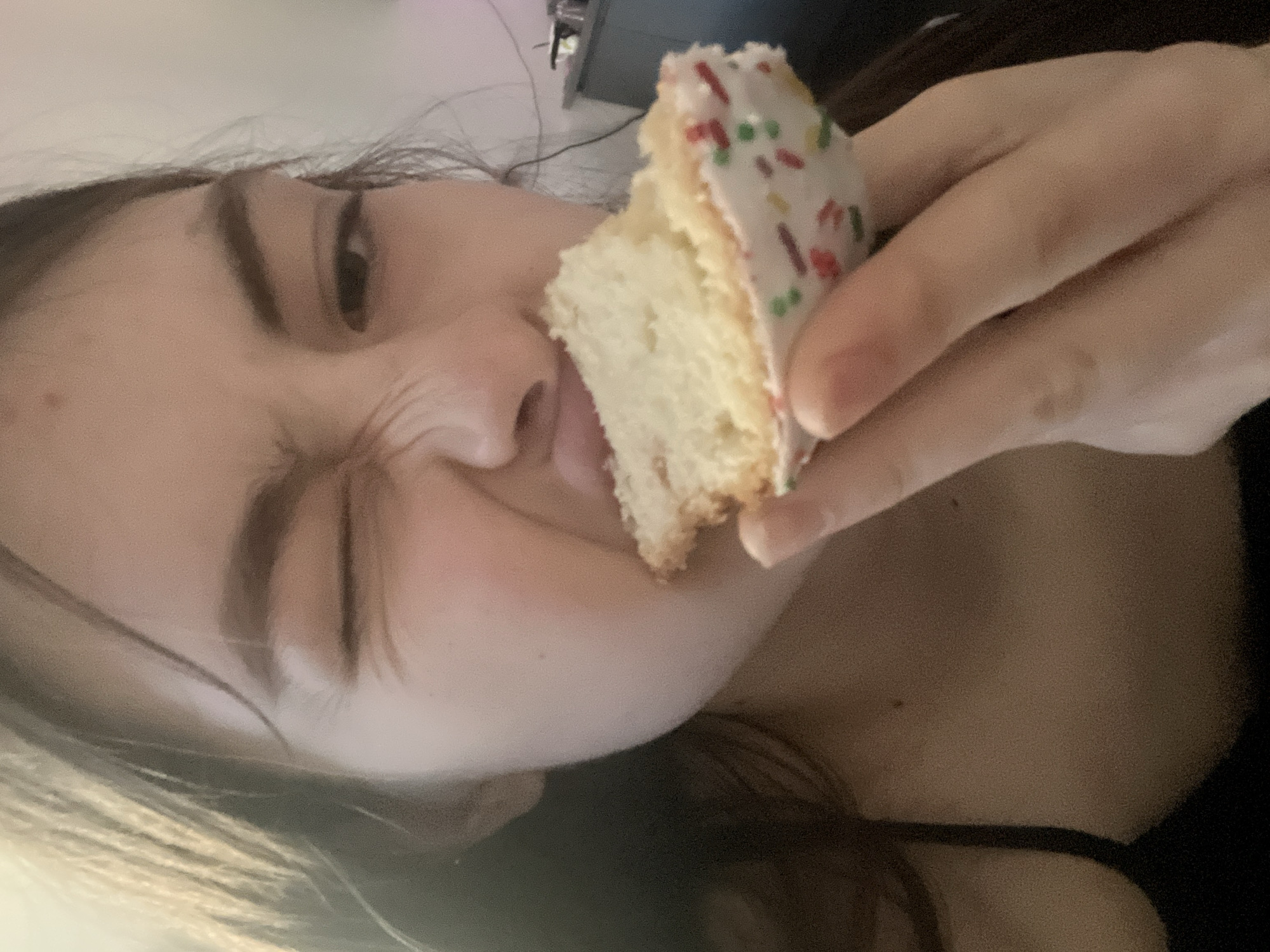 A fair-skinned woman is holding a piece of cake with white frosting and colored sprinkles to her mouth. She is grimacing or squinting, with her right eye closed. She has dark hair. She is wearing a black top. The background is blurry, but a gray cabinet is visible in the upper background. The location is indeterminable from the provided information; there is no indication this photo was taken in Saint Petersburg, Russia.