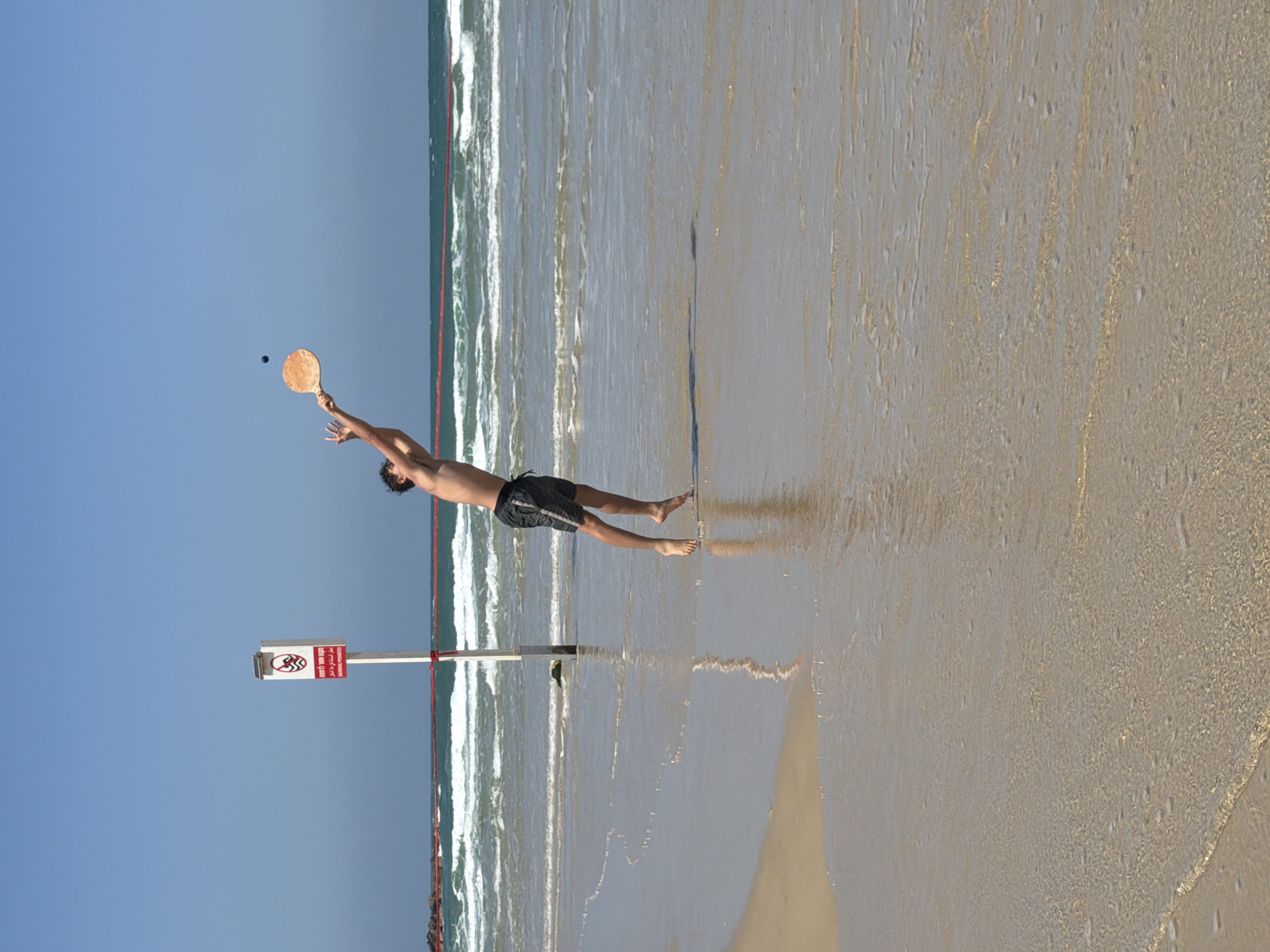 A man is leaping in the air to hit a ball with a paddle on a beach in Tel-Aviv, Israel. He is shirtless and wearing shorts. In the background, ocean waves crash on the shore. A sign with red markings is visible near the man. The sky is clear and blue.