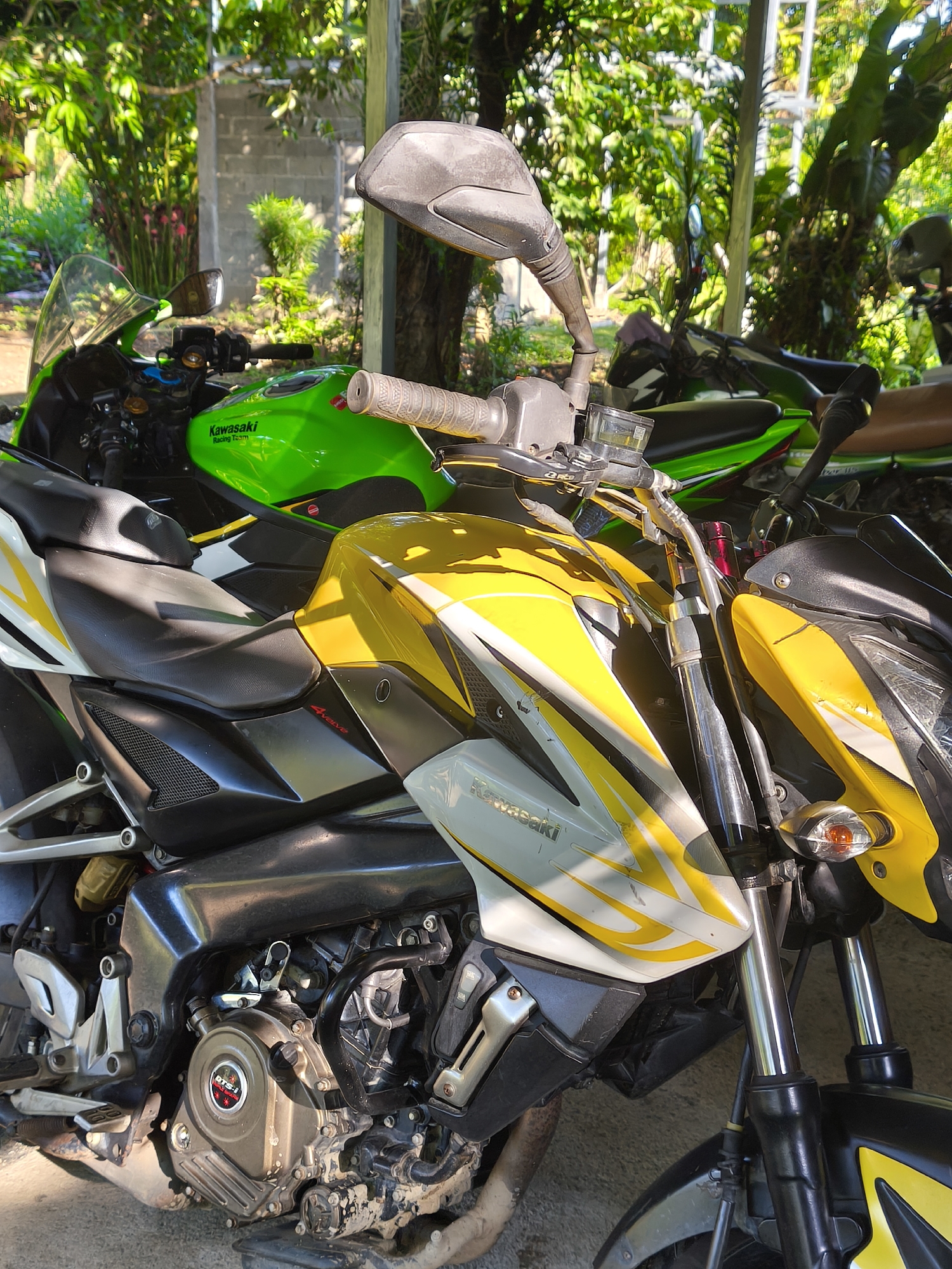 The image shows a close-up view of several parked motorcycles under a shaded structure. The primary motorcycle in the foreground is a yellow and white Kawasaki, with a black seat and engine. Behind it, partially obscured, is another Kawasaki motorcycle, predominantly green. More motorcycles can be seen in the background, adding to the overall scene of parked vehicles. The setting appears to be an outdoor garage or covered area, surrounded by lush greenery and trees. The location is described as Unknown, Unknown.