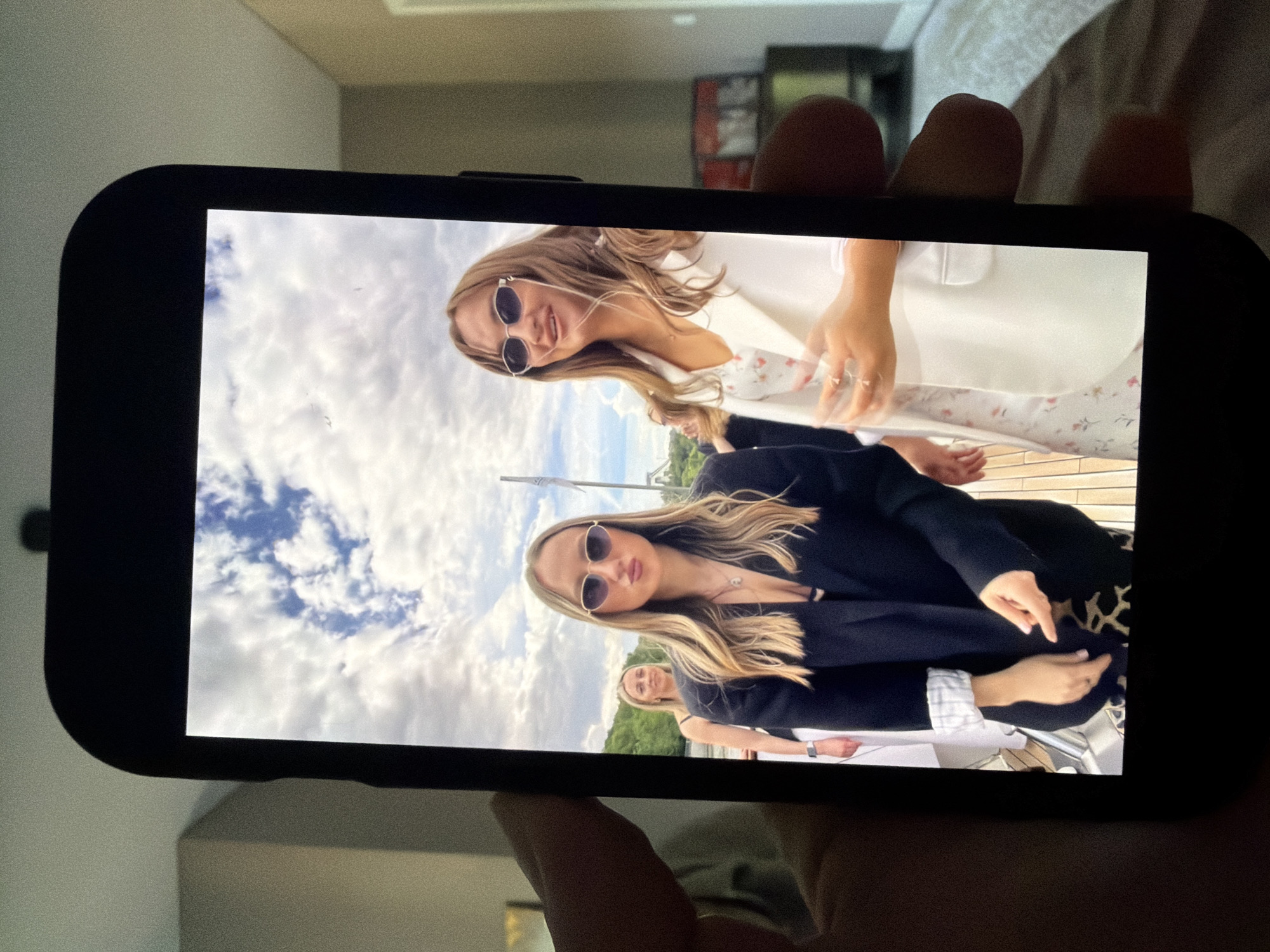 The image is a photograph of a smartphone screen displaying a picture taken outdoors, possibly on a boat. The photo on the screen shows three young adult females. The two in the foreground are positioned slightly off-center, with the one on the right slightly closer to the camera. Both are wearing sunglasses and appear to be posing for the photo. The female on the right is wearing a white blazer over a floral-patterned dress. The female on the left is wearing a black blazer over a dark top, and leopard-print pants. A third female, partially obscured, is visible behind them. The background features a cloudy sky and some greenery, suggesting an outdoor setting. The smartphone is held by a person whose hand is partially visible in the bottom left corner of the frame. The background of the original image, outside the smartphone screen, shows an indoor environment with furniture and a wall. There is no indication or information in the image that places the scene in Malinovka, Russia.