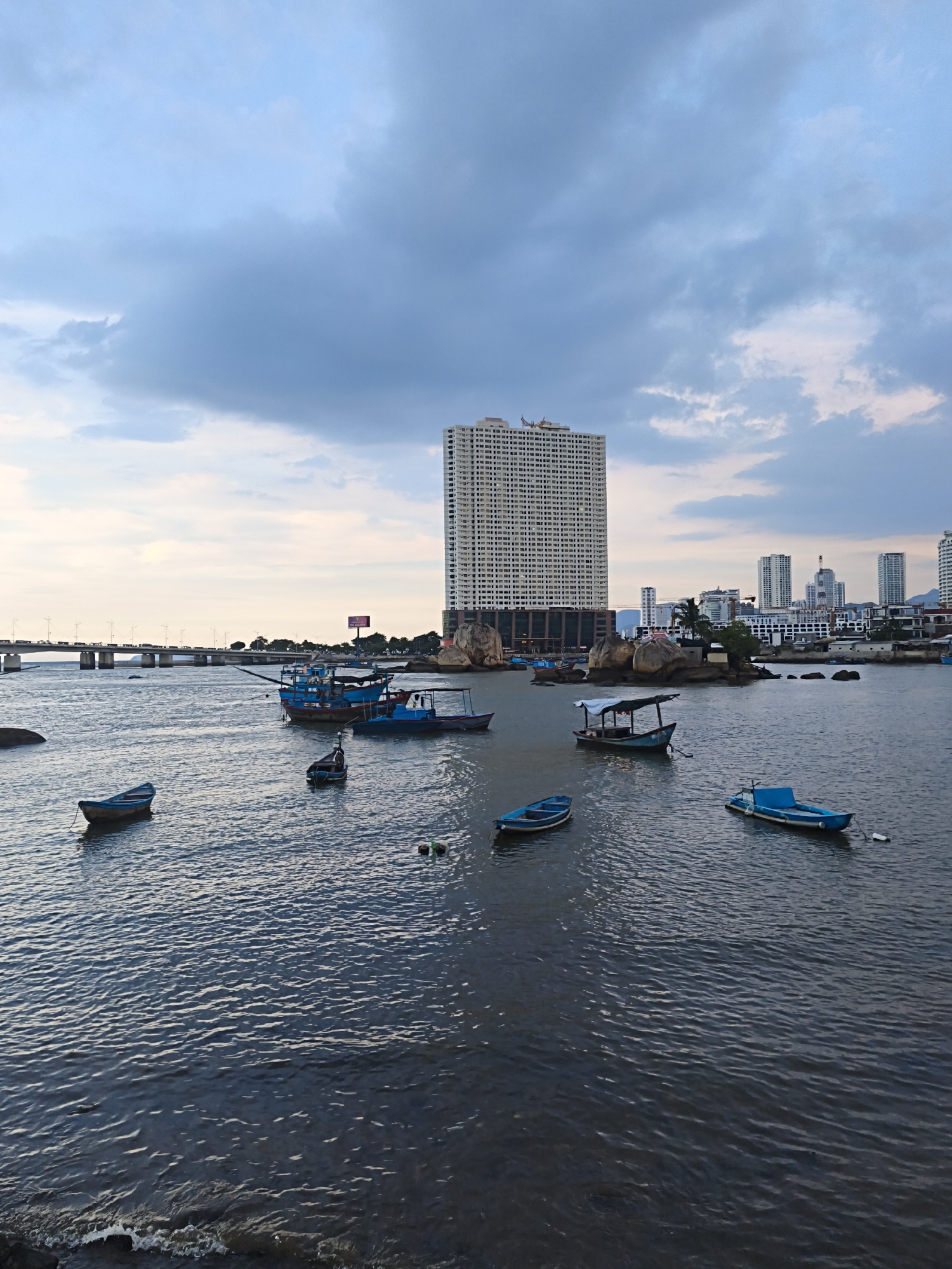 The image depicts a waterfront scene in the city of Nha Trang, Vietnam, under a cloudy sky. In the foreground, a body of water with gentle ripples is visible, with several small blue boats floating on its surface. One boat in the middleground is larger and appears to be a fishing vessel. In the background, a long bridge spans across the water, with traffic visible on it. A tall, white, multi-story building dominates the skyline, with other smaller buildings and structures situated near the shore. Rocky outcrops are interspersed with buildings along the coastline. The overall impression is of a coastal urban environment at dusk or early morning, given the muted lighting.