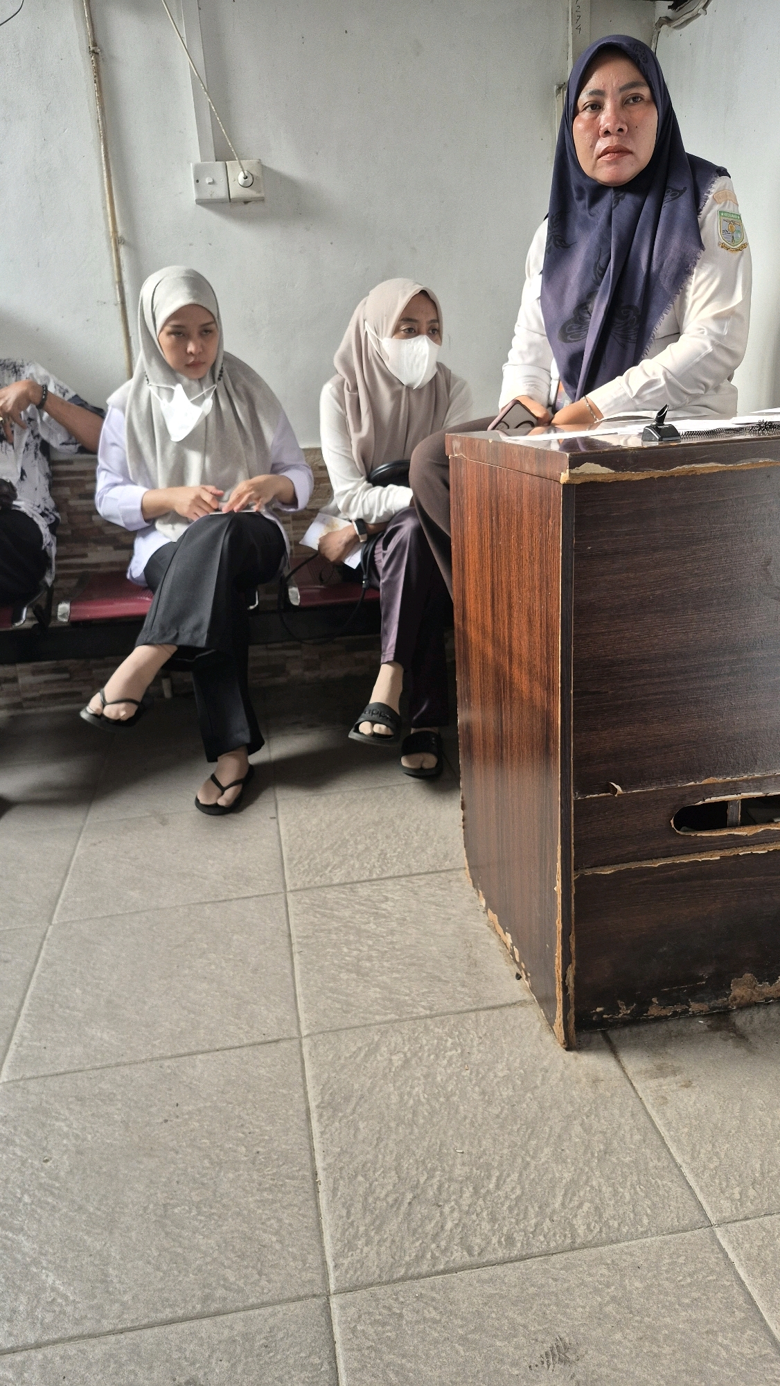 The image shows three individuals indoors, likely in Jambi, Indonesia, seated on a bench in front of a wooden desk. The person furthest to the right, wearing a dark blue hijab and a white long-sleeved shirt with a logo on the chest, is seated behind the desk and appears to be interacting with papers. Two other individuals, both wearing hijabs and masks, are seated on the bench to the left of the desk. One is wearing a light gray hijab and a white top with black pants, and the other is wearing a beige hijab and a white top with dark pants. The floor is tiled with a light gray material, and the wall behind the individuals is painted white with some electrical fixtures visible. The overall environment suggests a functional indoor space, possibly an office or waiting area, given the presence of a desk and seating.