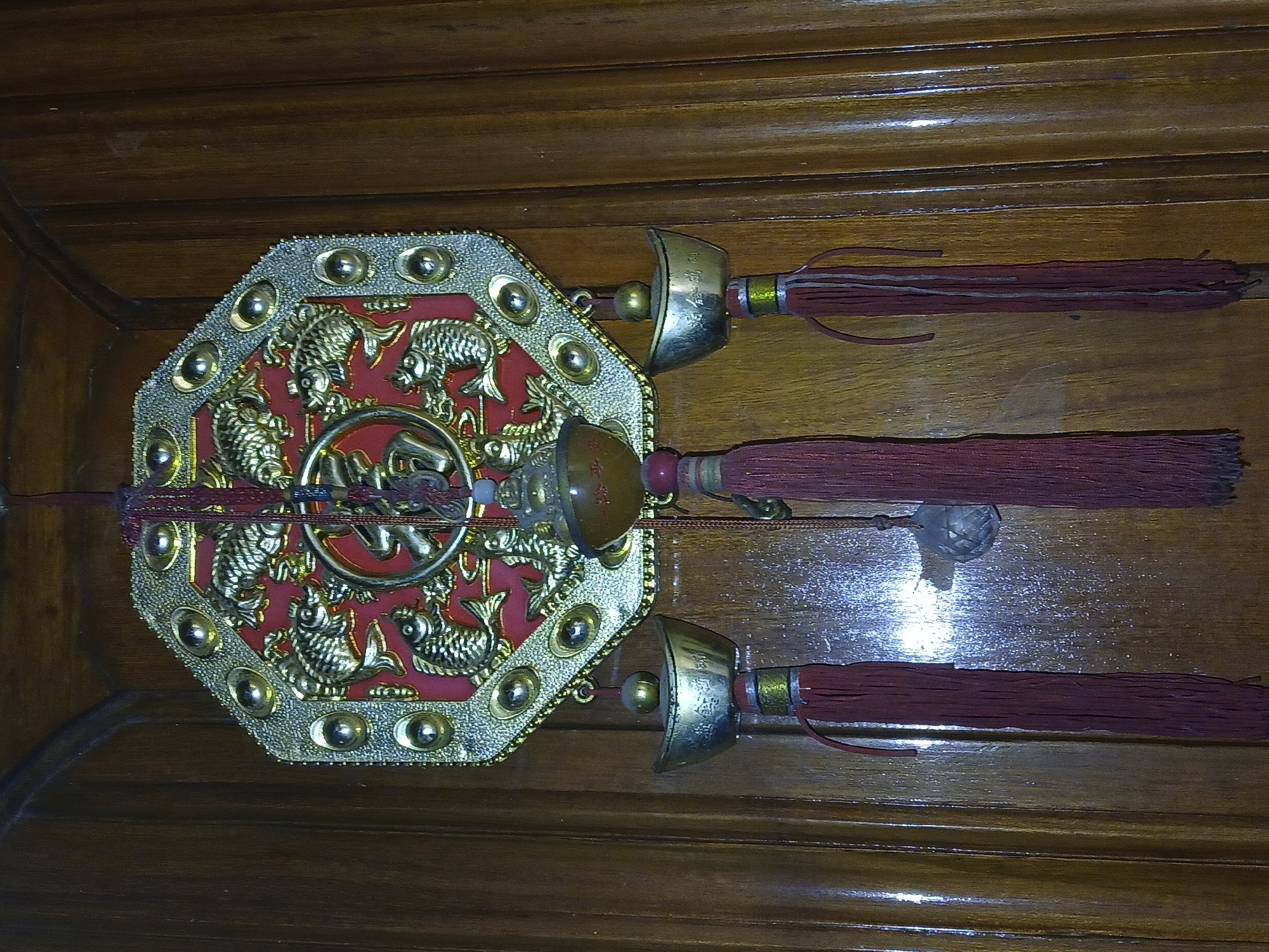 A decorative wall hanging lies on a wooden surface. The central element is an octagonal red and gold ornament with a pattern of fish. A golden ring with Chinese characters sits at the center. Three decorative attachments with short sword-like components and maroon tassels extend from the octagonal ornament. The wooden surface is stained and appears to be part of a piece of furniture. The image was taken indoors under artificial lighting. There is no evidence in the image to suggest it was taken in Bengaluru, India.