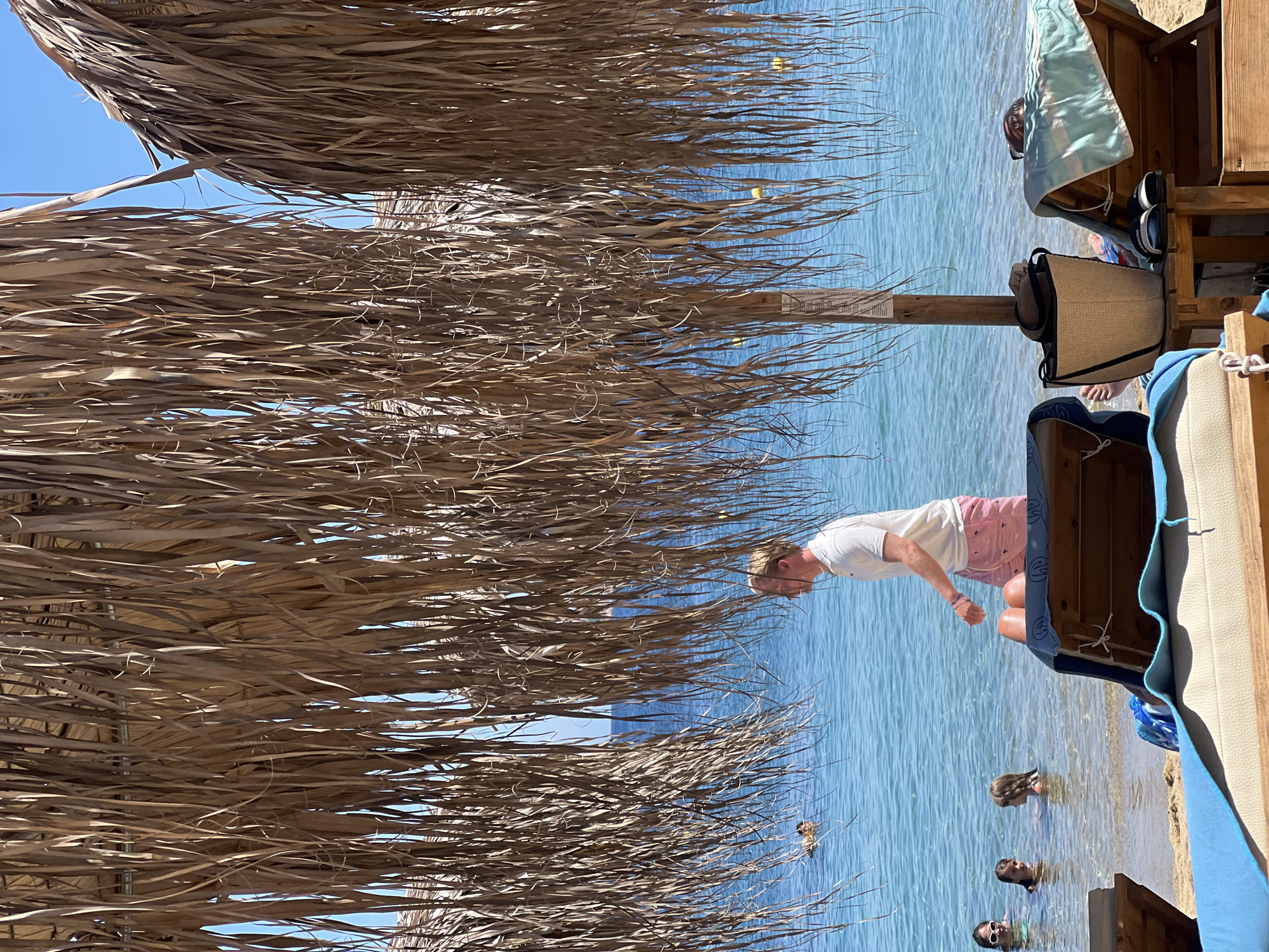 The image depicts a sun-drenched beach scene in Achladias, Greece, characterized by thatched umbrellas providing shade. In the foreground, two wooden lounge chairs are visible, one with a blue towel draped over it and another partially obscured. A woven beach bag rests on the wooden side table next to the second lounge chair. In the midground, a man in a white t-shirt and pink shorts walks towards the water, with his back to the camera. The shallow, clear blue water is populated by several children and a person swimming. The background reveals the vast expanse of the Aegean Sea under a clear blue sky. The overall ambiance is one of relaxation and summer vacation.