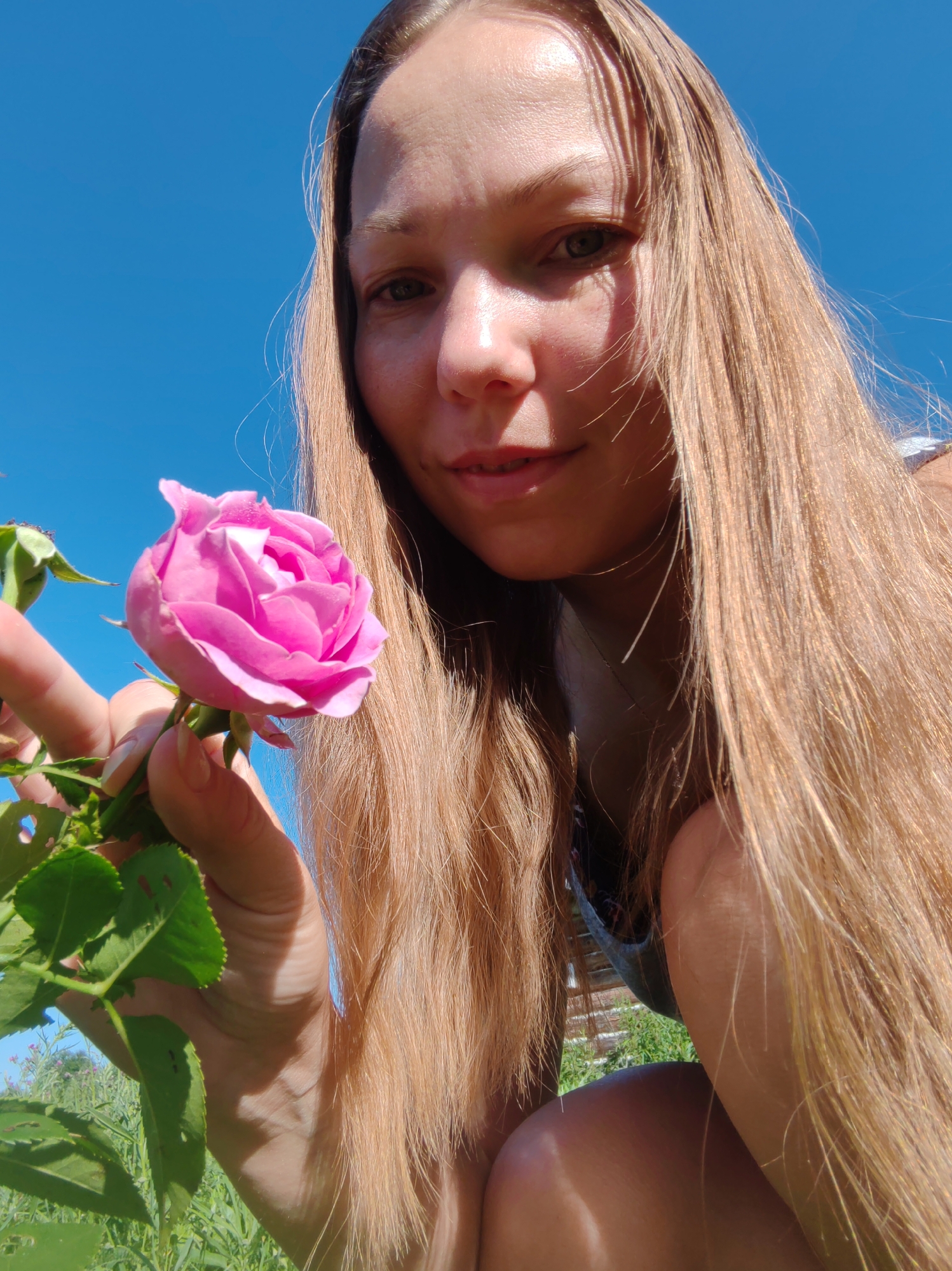 A young woman with long, blonde hair is shown from a low angle, holding a pink rose. She is outdoors, with a clear blue sky in the background. The image appears to be a selfie, with the woman looking towards the camera. The setting is natural and appears to be a garden or park, possibly in the city of Chuhlem, Russia, given the provided context. The woman is wearing a dark-colored garment.