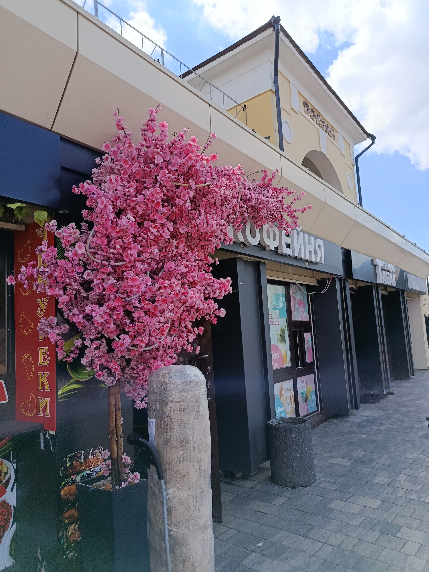 An outdoor shot captures a street scene in Orekhovo-Zuyevo, Russia, featuring a row of storefronts under a blue sky with scattered clouds. In the foreground, a faux cherry blossom tree with pink flowers is positioned in front of a dark-colored building facade. To the left of the tree, a red sign displays vertical text in Cyrillic. The building displays signage that reads "КОФЕЙНЯ" (Coffee Shop) and "ПРАВДА ТАБАК" (Pravda Tobacco). Windows of the coffee shop show colorful advertisements for drinks like iced matcha and tea. A cylindrical trash bin stands near the entrance. A light-colored, cylindrical object wrapped in plastic is situated to the right of the tree, with a black handle or hose attached. The ground is paved with grey interlocking bricks. In the background, the upper section of a building features a light yellow facade with arched windows and decorative elements, and text that reads "ВОКЗАЛ" (Station).