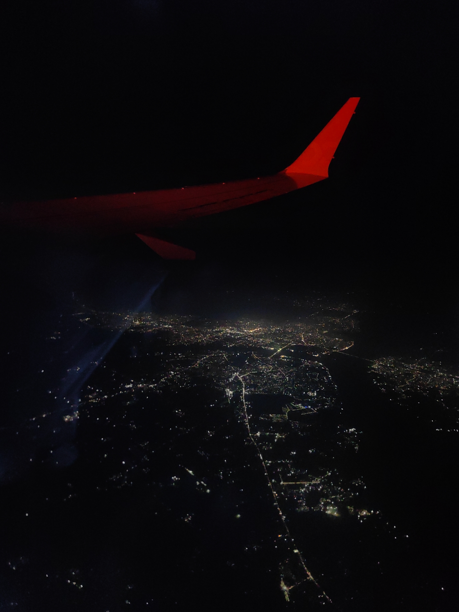 The image is an aerial view taken at night from an airplane window, showing the wingtip of the aircraft illuminated in red. Below, a city is visible, illuminated by numerous lights. The lights are scattered, suggesting a densely populated area. A body of water, likely a river or lake, appears in the lower half of the image, with lights reflecting on its surface. Several bright beams of light, possibly from searchlights or high-powered lamps, are visible cutting through the darkness over the city. The presence of these lights indicates activity within the city at night. The exact location within Tendui Ta. Khurd, India cannot be determined from the image alone. There are no people or distinct objects visible on the ground.