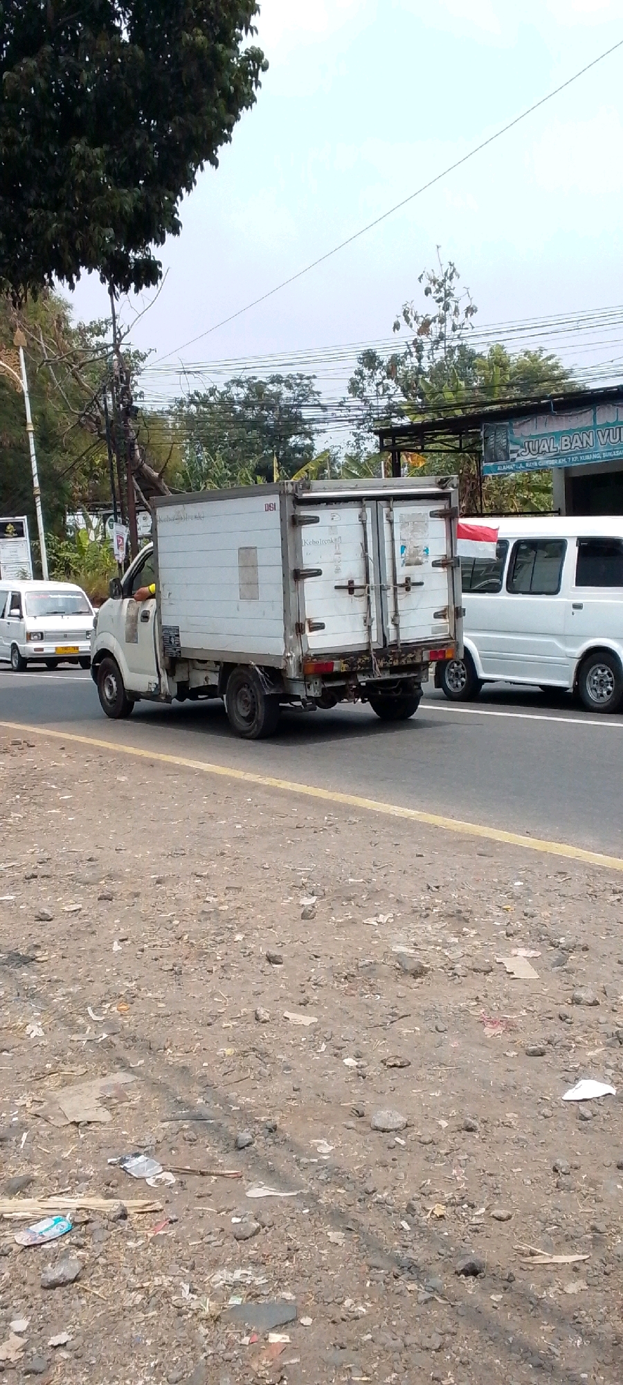 In Cilaku, Indonesia, a white box truck is captured from a side-rear perspective on a paved road. The truck, appearing to be a delivery vehicle, has its rear doors closed and a white, rectangular cargo box. Its wheels are black, and the vehicle shows signs of wear and tear, with some rust visible on the rear bumper and undercarriage.
To the left of the box truck, a white minivan is also on the road, facing the same direction. This minivan has tinted windows and chrome rims. In the background, a variety of trees and vegetation line the roadside, interspersed with power lines and utility poles. A sign, partially visible, reads "JUAL BAN VULCANIZE" (Selling Vulcanized Tires), suggesting a commercial area. The road surface is asphalt, with a yellow line marking the lane division. The foreground consists of a dirt and gravel shoulder, littered with debris including plastic wrappers, paper, and small stones. The sky is overcast with a hazy appearance.