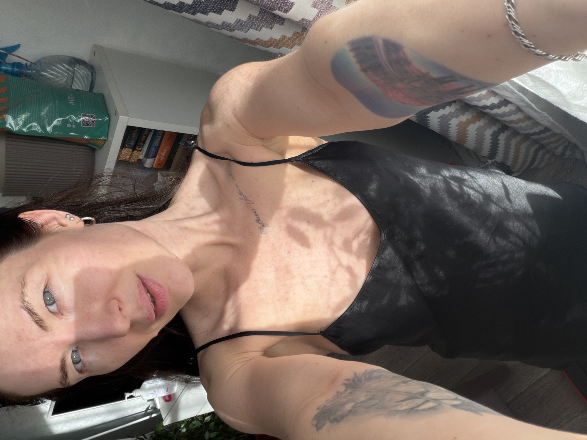 A woman with pale skin and blue eyes is posing in a selfie, wearing a black tank top. She has tattoos on both arms and some text on her upper chest. She also has multiple piercings in her left ear. The background includes a shelf with books and a green package. The location seems to be indoors. There is no evidence to determine that the location is in the city of Perm, Russia.