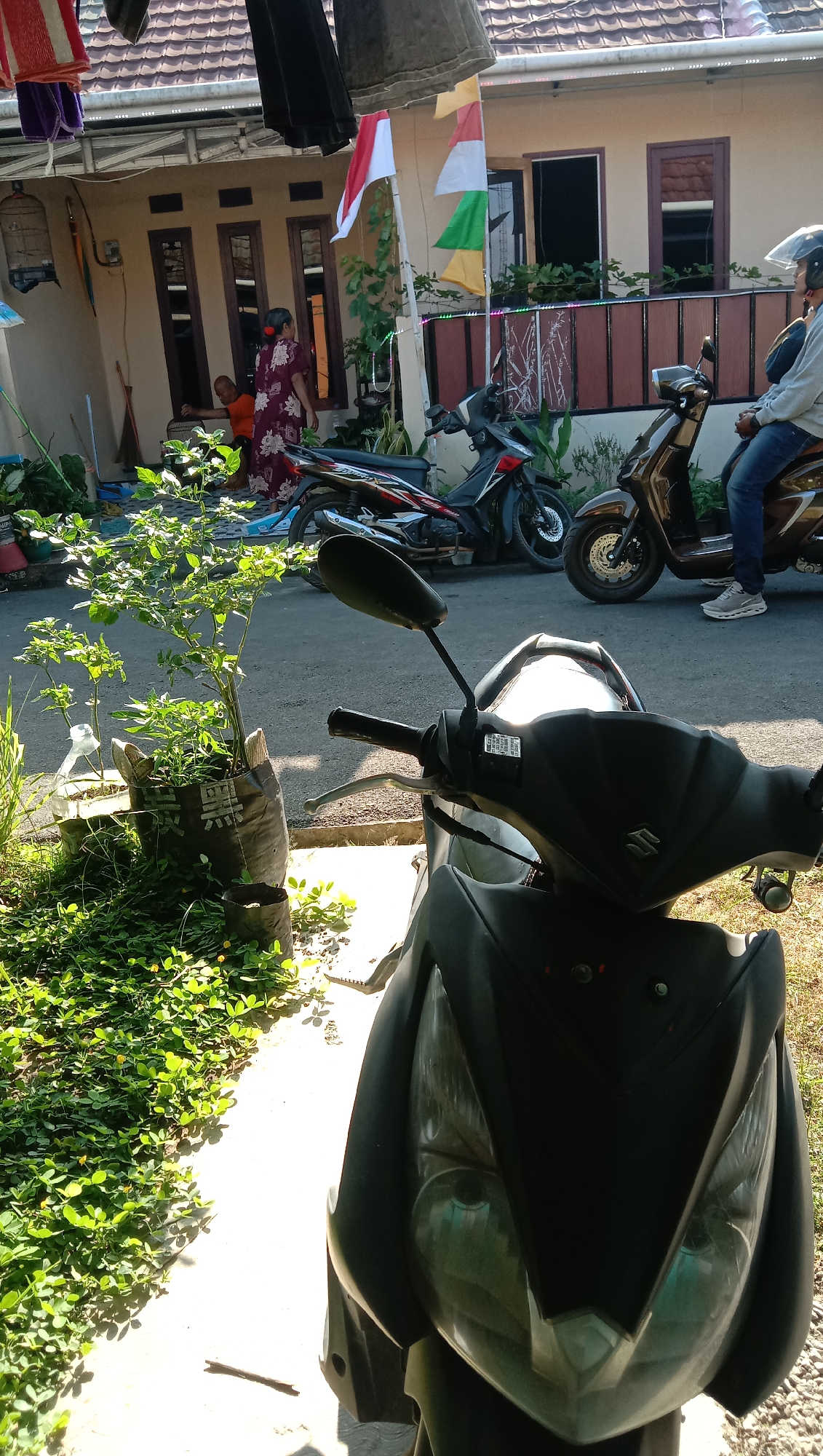 In Karangmangu, Indonesia, on a sunlit day, a scene unfolds in front of a residential building. Two motorcycles are parked on the asphalt road. The foreground is dominated by the front of a black scooter, its handlebars and headlight clearly visible. To the left of the scooter, a potted plant with green leaves sits on a paved area, surrounded by low-lying green foliage. Behind the motorcycles, the facade of a house is visible, with three windows and a dark-colored roof. A woman in a patterned dress stands near the entrance, looking towards a small child sitting on the ground. Several Indonesian flags, red and white, are attached to poles in front of the house, fluttering in the breeze. Laundry hangs on a line above the scene, adding to the domestic atmosphere.
