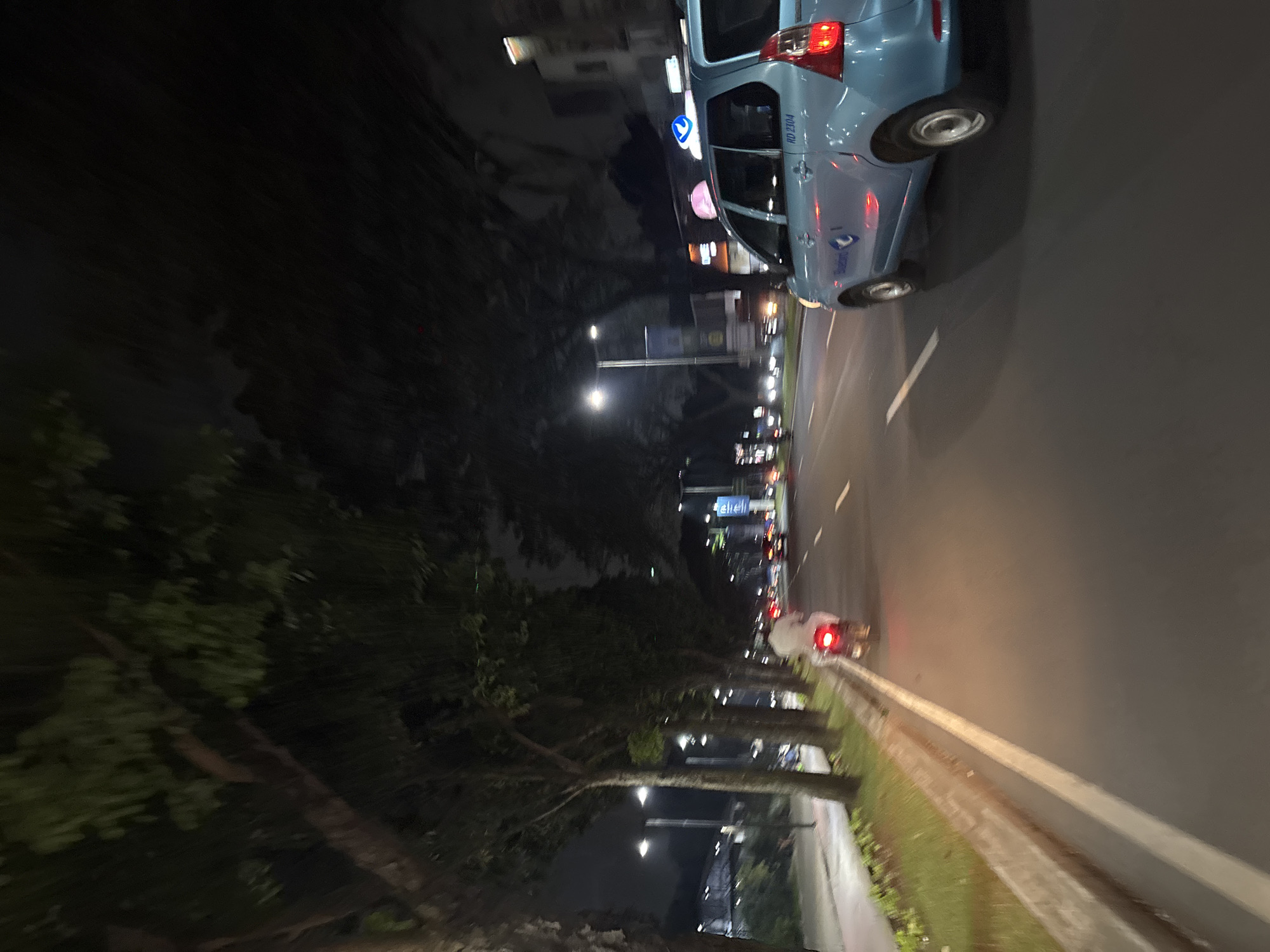 This media file captures a night-time street scene, likely taken from a moving vehicle in South Tangerang, Indonesia. The image is composed of a multi-lane road on the right, bordered by a concrete barrier and a grassy median to its left.
A light blue car, possibly a taxi, is prominently featured in the middle-right, its red taillights illuminated. Visible on its side is the text "HD 2304" and a logo resembling the Blue Bird taxi company, with "Bluebird" faintly readable below it. Further down the road, other vehicle lights indicate ongoing traffic.
To the left of the road, a dense line of trees with significant motion blur suggests the photograph was captured while moving. Streetlights illuminate parts of the road and the foliage. In the distant background, beyond the trees, faint lights from buildings and a blurry blue sign are visible, with text that could possibly be "Citilink," an Indonesian airline.
The scene conveys the atmosphere of an active urban or suburban environment at night, characterized by artificial lighting and moving vehicles. No specific individuals are discernible, and the weather appears clear.