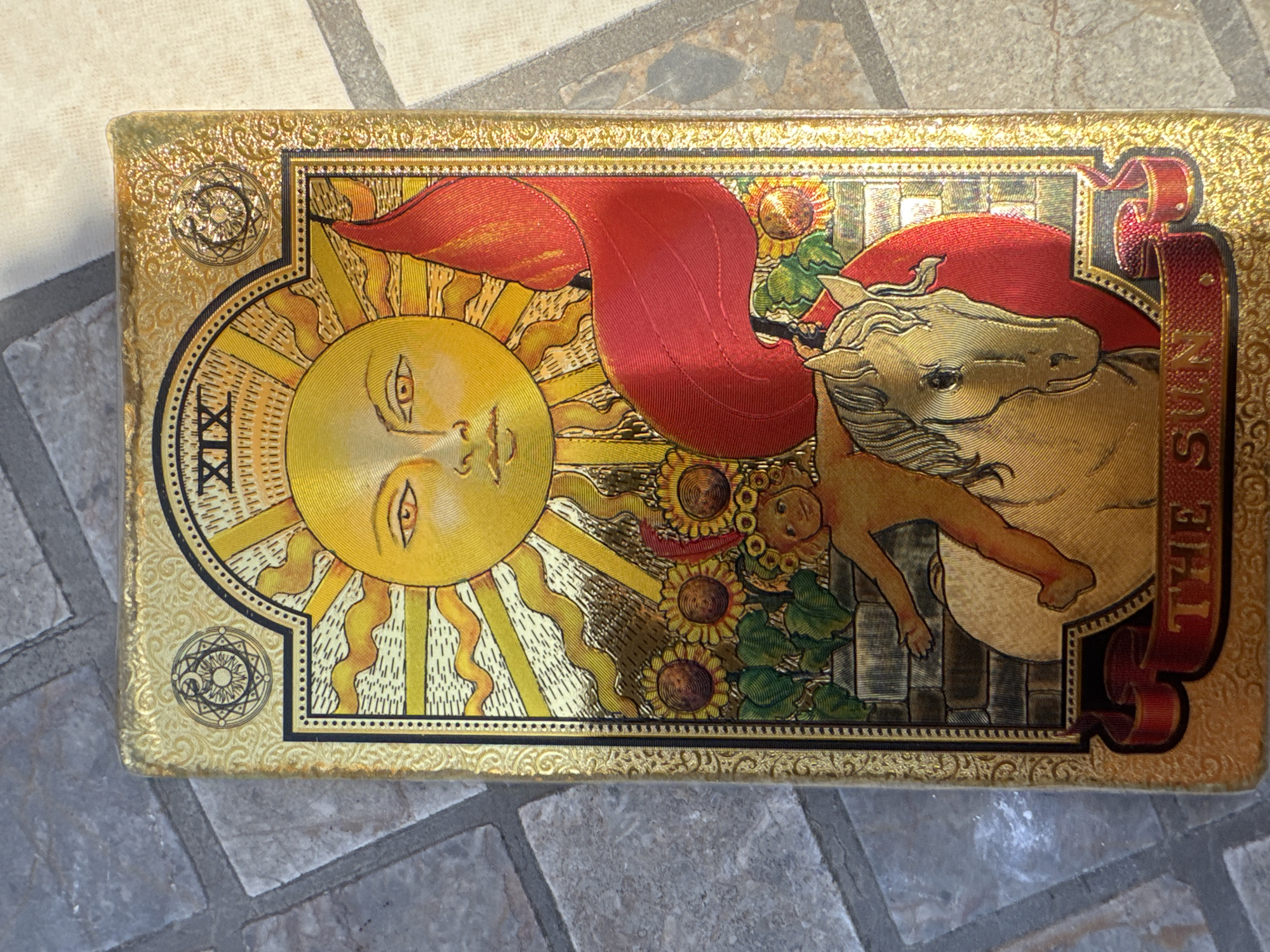 The image shows a rectangular object, likely a tarot card box, placed on a surface resembling tile. The box is decorated with an illustration of the "The Sun" tarot card. The design includes a sun with a face, sunflowers, a naked child, and a white horse. The text "XIX" is present, likely indicating the card's number in the Major Arcana. The words "THE SUN" are also visible in a ribbon-like design. The color scheme is predominantly gold, yellow, and red. The edges of the box are ornate with decorative patterns. There is no indication of location in Đà Nẵng, Vietnam.