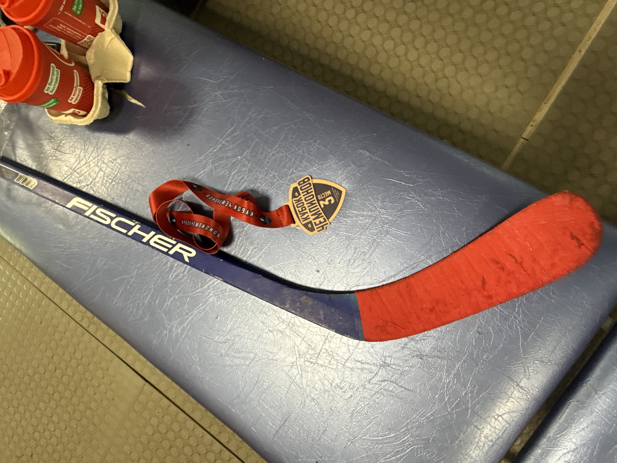 The image shows a Fischer brand hockey stick, with its blade wrapped in red tape. Next to it is a lanyard with a medal that reads "KYBOK-CHEMPIOHOB", indicating a hockey championship. The medal also denotes "3 MECTO", implying a third-place achievement. The objects rest on a blue padded surface, possibly a bench. In the upper left corner, there's a cardboard drink carrier containing multiple red cups. Text in Russian is visible on the cups, which includes "За воздушной пенкой" (for airy foam) and "За быстрой доставкой" (for fast delivery). The background appears to show a tiled floor or wall. Given the Cyrillic script and the name of the championship, it is likely that this scene is in Russia, potentially in the city of Izhevsk.