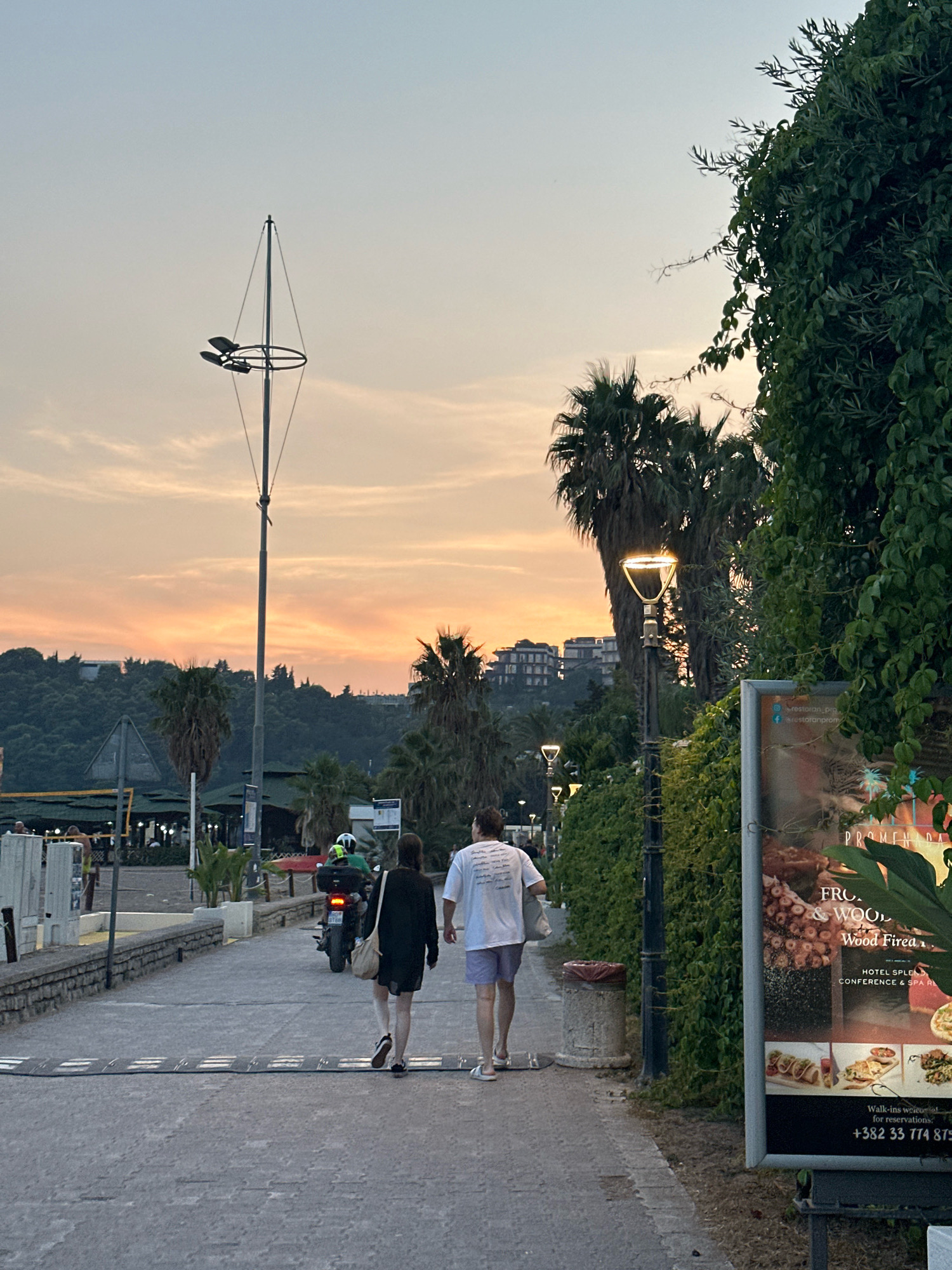 The image captures a scenic promenade in Boreti, Montenegro, at dusk. The sky displays a warm, gradient of orange and pink hues, indicating the setting sun. A tall, modern lamppost with multiple lights stands prominently in the foreground, casting a soft glow.
The promenade itself is paved with textured material and features speed bumps. To the right, a dense, lush green hedge lines the path, with a tall, modern lamppost integrated into it. A large advertising board is affixed to the hedge, displaying text and images of food and flowers.
Two individuals are walking away from the camera on the promenade. The person on the left is wearing a dark dress and carrying a bag over their shoulder. The person on the right is wearing a white t-shirt and light-colored shorts. Further ahead, a person on a motorcycle, wearing a helmet, is also moving along the promenade.
In the background, palm trees are visible, characteristic of a coastal environment. Beyond the trees, a hillside is covered with dense vegetation and dotted with buildings. Other streetlights are visible in the distance, illuminating the path.
To the left of the promenade, there is a stone wall separating it from an area that appears to be a beach or waterfront. Beach umbrellas and seating areas are visible, along with a volleyball net. People can be seen in the distance near the waterfront. The overall scene suggests a relaxed evening atmosphere by the sea.