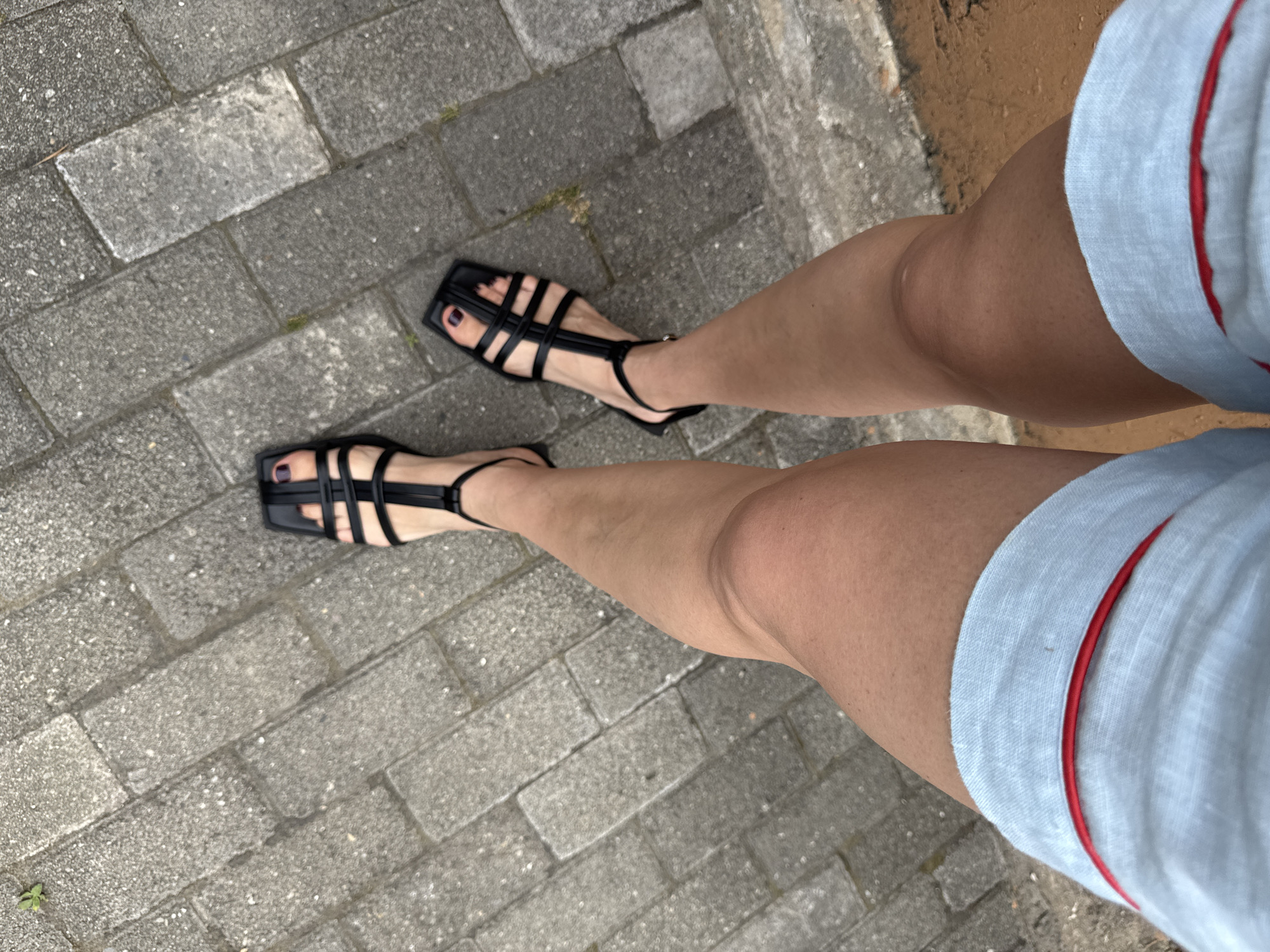 This is a first-person, over-the-shoulder view of a person's legs and feet, standing on a grey brick pathway. The person is wearing black, strappy, square-toed sandals with dark-colored nail polish on their toes. They are also wearing light blue shorts with a red trim. The pathway appears to be outdoors, possibly in a park or urban setting, as there are small patches of grass and dirt visible between the bricks. No specific landmarks or details suggesting the city of Sochi, Russia are present in the image.