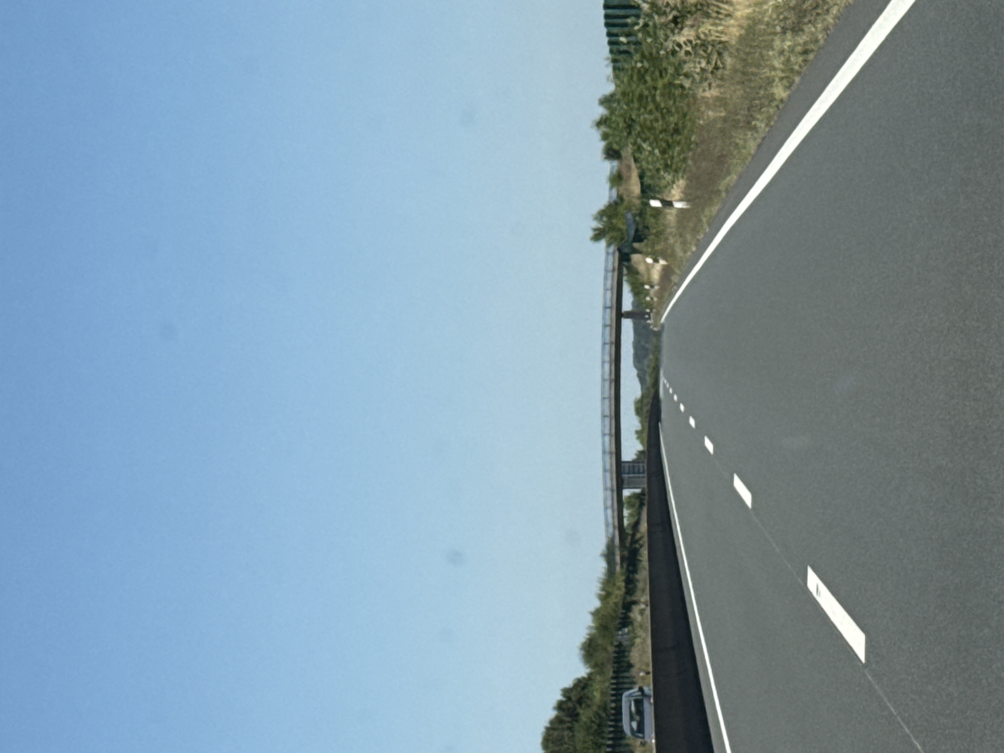 The image depicts a highway in Bad Sachsa, Germany, on a clear, sunny day. The road has two lanes, marked by white lines, and a concrete barrier on the left side. A pedestrian bridge spans the highway in the distance. Vegetation, including trees and dry grass, lines the sides of the road, with some black and white striped posts visible along the embankment. A dark-colored car is parked on the shoulder of the highway on the left side. The sky is a bright, cloudless blue.