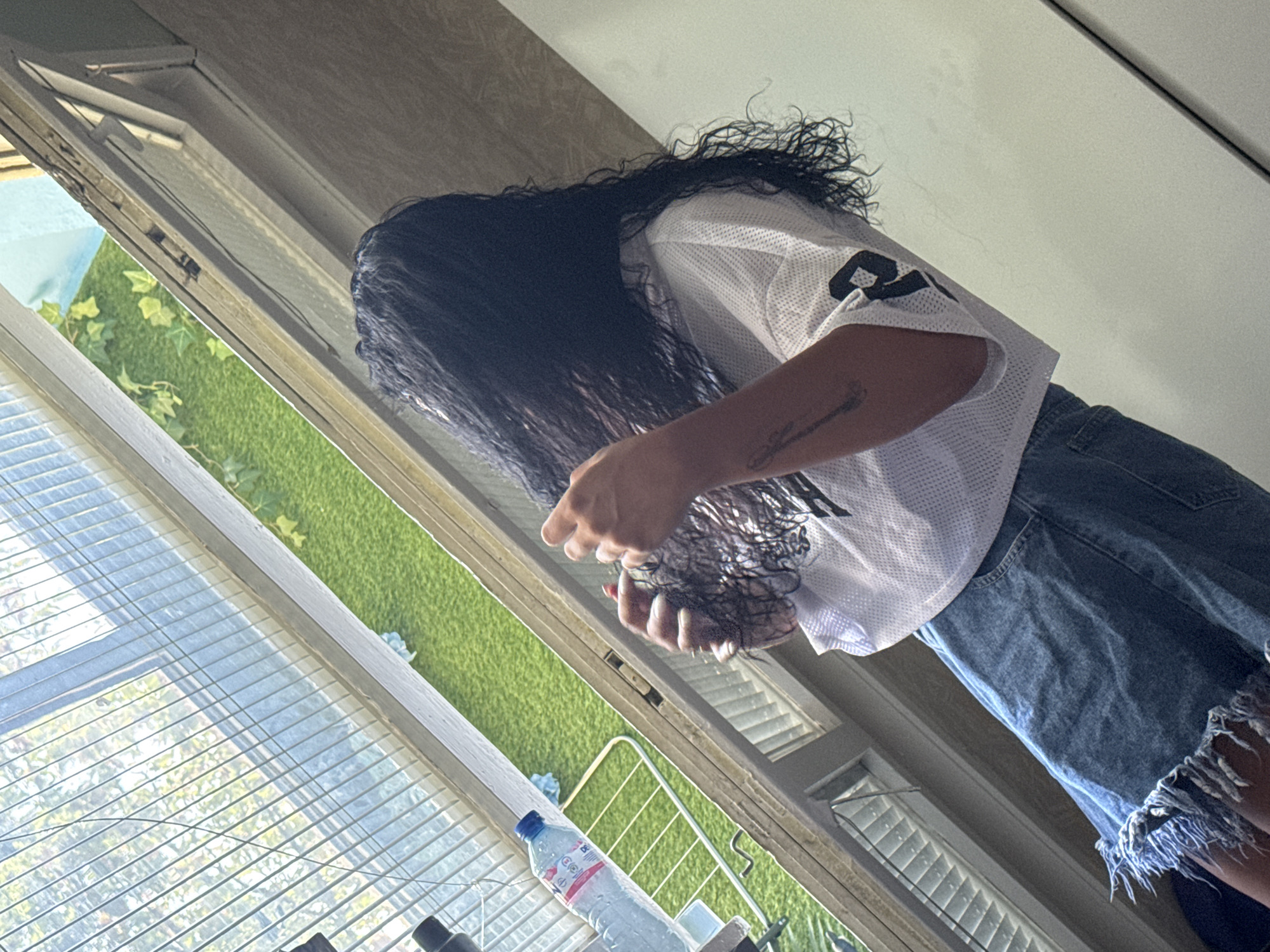 The image captures a person with long, dark, wet hair, wearing a white mesh jersey with black numbering and distressed denim shorts. The person is leaning forward, appearing to be styling or drying their hair, with their hands near their head. They have a tattoo on their left forearm. The scene is illuminated by natural light streaming in from a window. Outside the window, a bright green, artificial grass-covered surface with some ivy-like plants is visible, suggesting an outdoor or enclosed garden space. Inside, blinds cover part of the window, and a bottle of water is placed on a surface in the foreground. The perspective is angled, looking upwards at the subject. The presence of artificial grass and the specific angle of the light might indicate a location in Sofia, Bulgaria, potentially a residential area or a place with a curated outdoor space, though no specific landmarks confirm this.