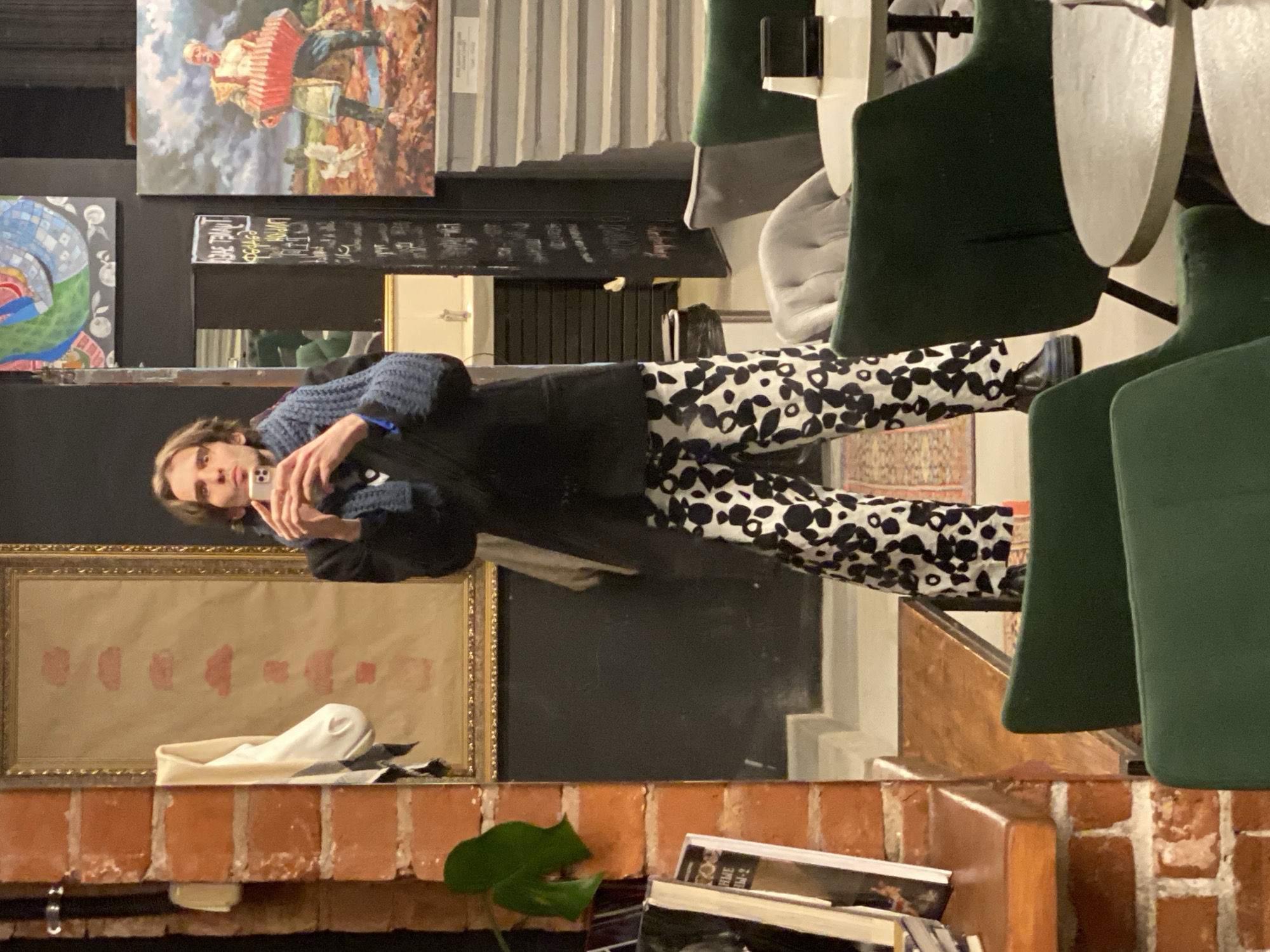 A person is reflected in a mirror. The mirror shows their upper torso and legs, they are wearing a blue sweater and black jacket, with black and white patterned pants. They are holding a cell phone in their hand. Above the mirror are paintings on the wall. Below the mirror is a brick ledge with books and a plant. A series of green chairs is on the right. Based on the location shown in the image and metadata, this location is in Moscow, Russia.