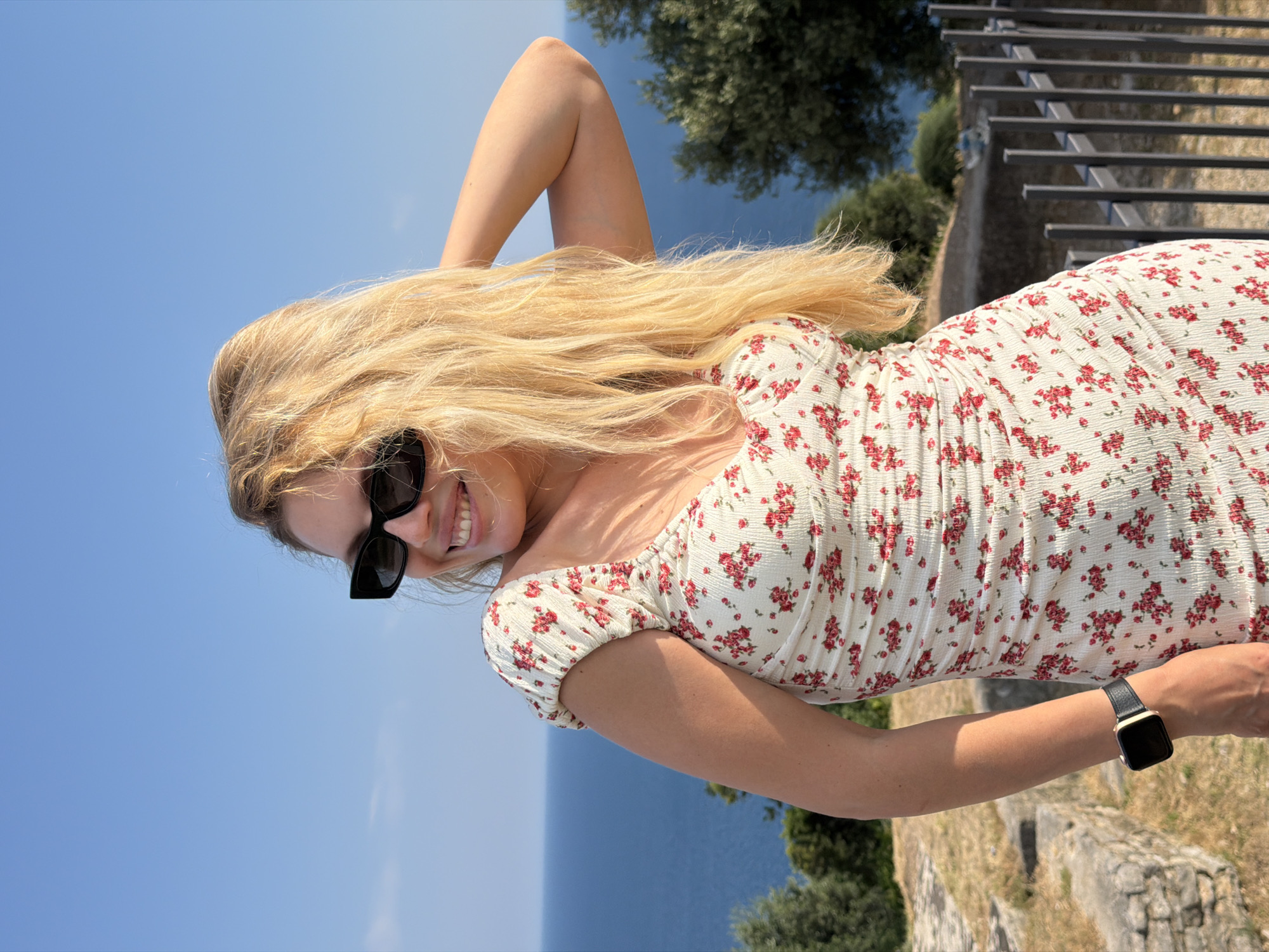 A blonde woman wearing a white floral dress and sunglasses poses for a photo outdoors. She is standing in front of a blue ocean and sky, with some trees and ruins visible in the background. The woman has her hand in her hair and is smiling. She is also wearing a watch on her left wrist. The scene appears to be located in Budva, Montenegro, given the architecture and vegetation.