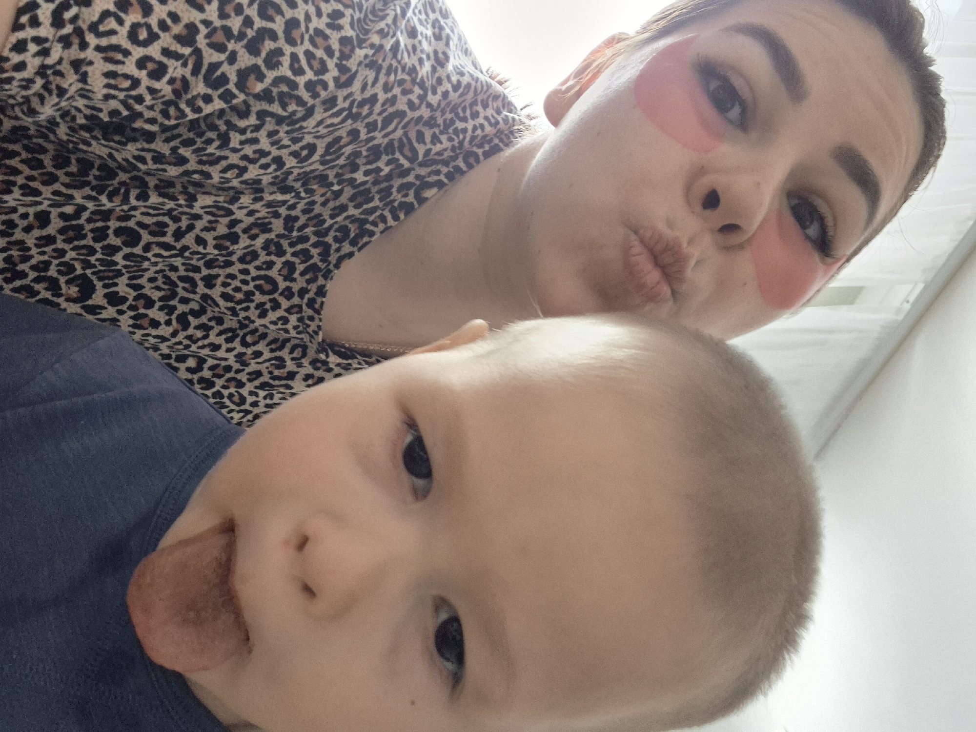A selfie taken from above shows a woman and a baby. The woman has pink eye patches and is pouting her lips. She wears a leopard print top. The baby, wearing a blue top, sticks out its tongue. The location is unknown, and there is no indication that the image was taken in Tobolsk, Russia. The background appears to be a white wall.