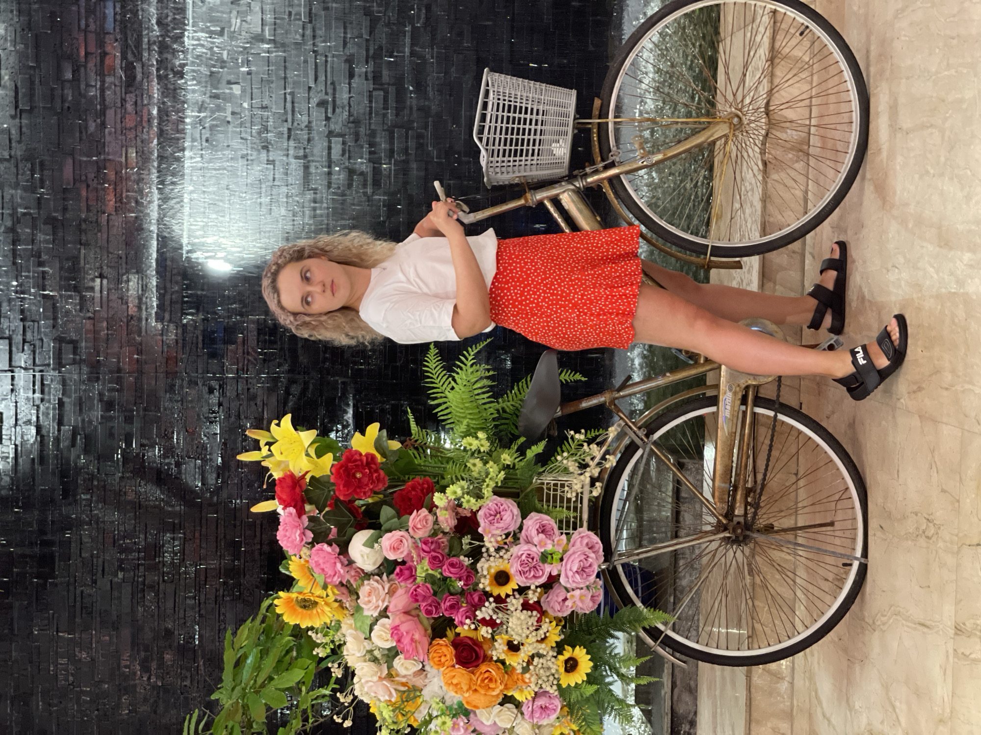 A young, fair-skinned woman stands next to a gold-colored bicycle in Nha Trang Ward, Vietnam. The woman has curly blonde hair, and wears a white t-shirt, a short red skirt with white polka dots, and black sandals. The bicycle is adorned with a basket and a large bouquet of flowers including sunflowers, roses, lilies, and other flowers of varied colors. The background includes a textured black brick-like wall with a water feature, and a light-colored tiled floor.