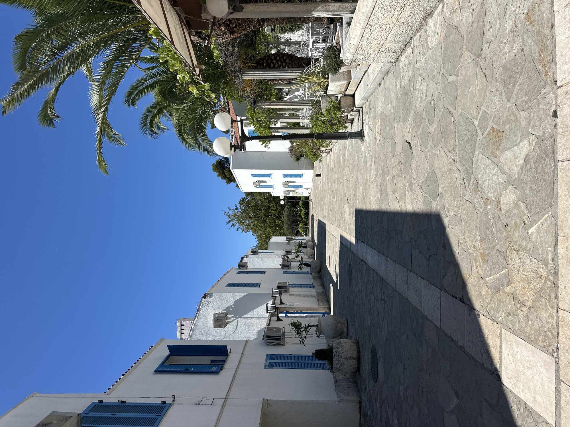 The image shows a narrow, sunlit stone street in Budva, Montenegro. The street is lined with white buildings featuring blue shutters and air conditioning units. A row of street lamps runs along the street, casting a shadow. Palm trees are visible in the upper left corner. A stone walkway extends along the right side of the street, leading towards a pergola-covered area with white chairs. The sky is clear and blue.