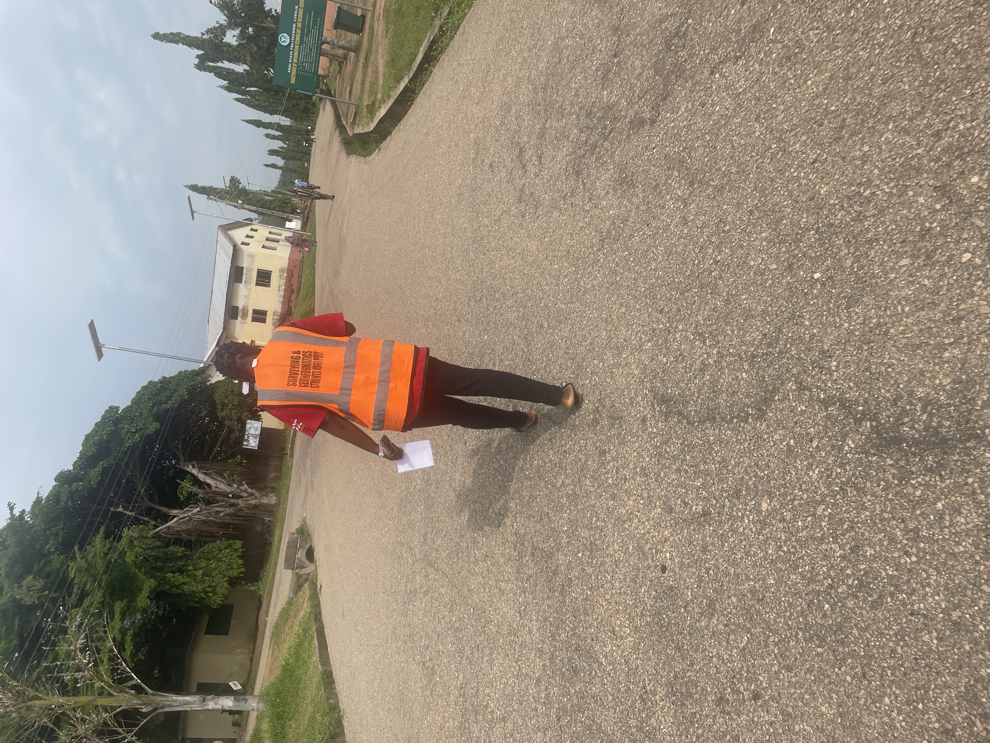 In Lokoja, Nigeria, a person wearing a bright orange safety vest with text that reads "SURVEYING & GEODETIC INFORMATICS STUDENTS KOGI POLY" walks away from the camera on a paved road. The person is holding a white piece of paper in their left hand. The road curves to the right and is lined with green grass and trees. In the background, there are buildings and a sign that says "KOGI STATE POLYTECHNIC, LOKOJA." The sky is overcast.