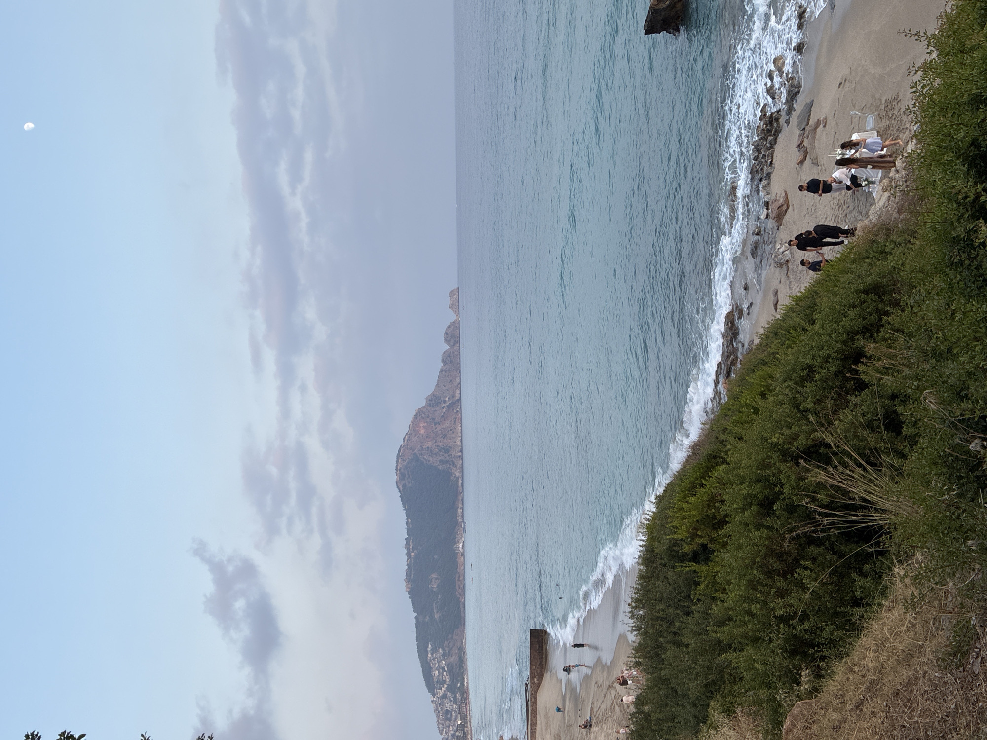 The image shows a coastal scene with a beach in the foreground and a large hill with buildings and a fort in the background. The sea is calm with gentle waves breaking on the shore. The sky is clear with some scattered clouds and the moon is visible.
In the foreground, there is a group of people on the beach. Some are standing and talking, while others are seated at a table that appears to be set up for an event. The table is covered with a white tablecloth and has candles and place settings. There are chairs around the table, and some people are standing near it.
Further down the beach, more people are visible, some walking and others wading in the water. The beach is sandy with some rocks and pebbles. Dense green vegetation covers a hillside in the lower part of the image, obscuring the view of the ground there.
In the background, a prominent hill overlooks the sea and the coastline. The hill is covered with trees and vegetation, and there are many buildings clustered on its slopes, indicating a town or city. A fortified structure is visible at the peak of the hill. The water extends to the horizon, where the sky transitions to a lighter shade of blue. A large rock formation is also present in the water near the shore. The overall impression is a scenic coastal location, likely in or near Alanya, Turkey, given the distinctive topography and architecture.