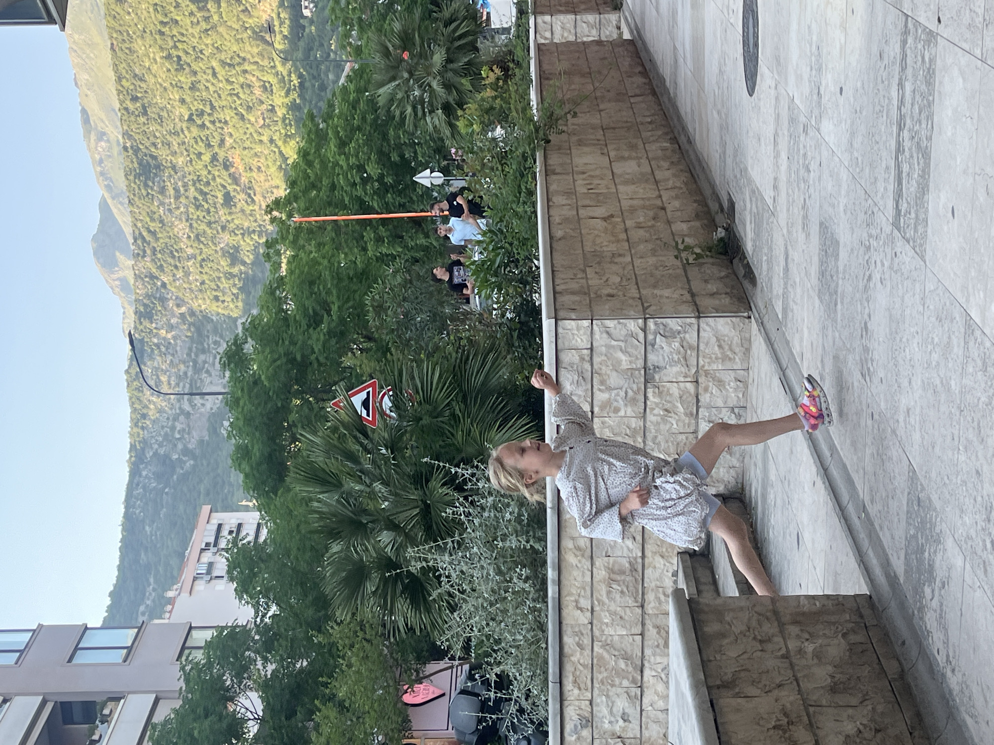 The photograph captures a young girl standing on a ledge in Budva, Montenegro, with a mountain range visible in the background. The girl is wearing a patterned dress and brightly colored sneakers, her arm raised as if pointing at something out of frame.
In the background, a traffic sign is partially obscured by foliage, and several people are gathered nearby an orange boom. An apartment building is visible to the left. The scene suggests an urban environment near natural landscapes, with the architecture, vegetation, and mountain providing a sense of location.