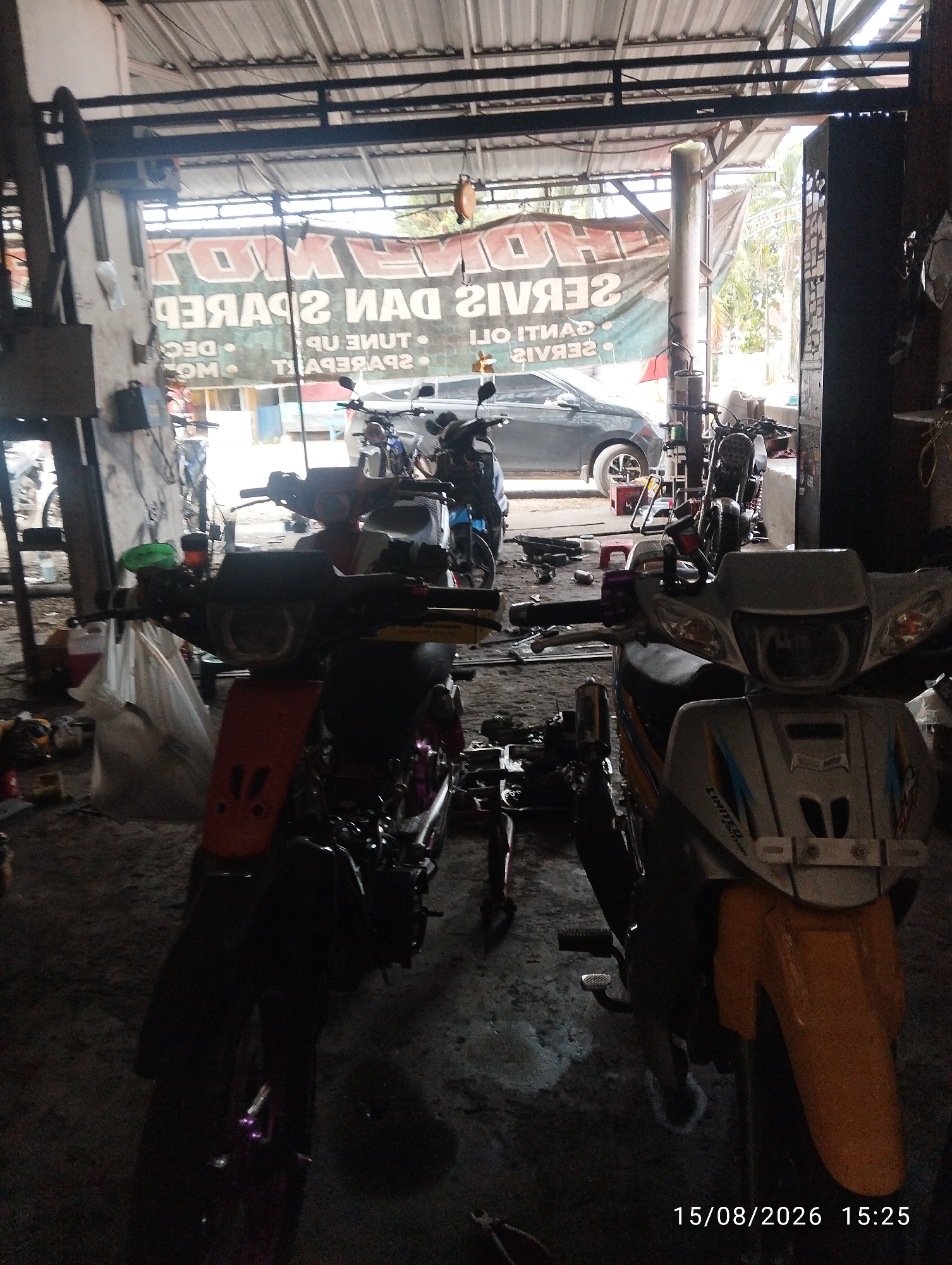 The image depicts the interior of a motorcycle repair shop in Tegal Rejo, Indonesia. Two motorcycles are prominently displayed in the foreground, one orange and black, and the other silver and yellow. In the background, a dark grey car is parked, and other motorcycles and shop equipment are visible. A banner advertising "SERVIS MOTOR DAN SPAREPART" (Motorcycle Service and Spare Parts) is hung on the wall. The environment appears to be a typical workshop with concrete flooring and a metal roof. The timestamp "15/08/2026 15:25" is visible in the bottom right corner of the image.