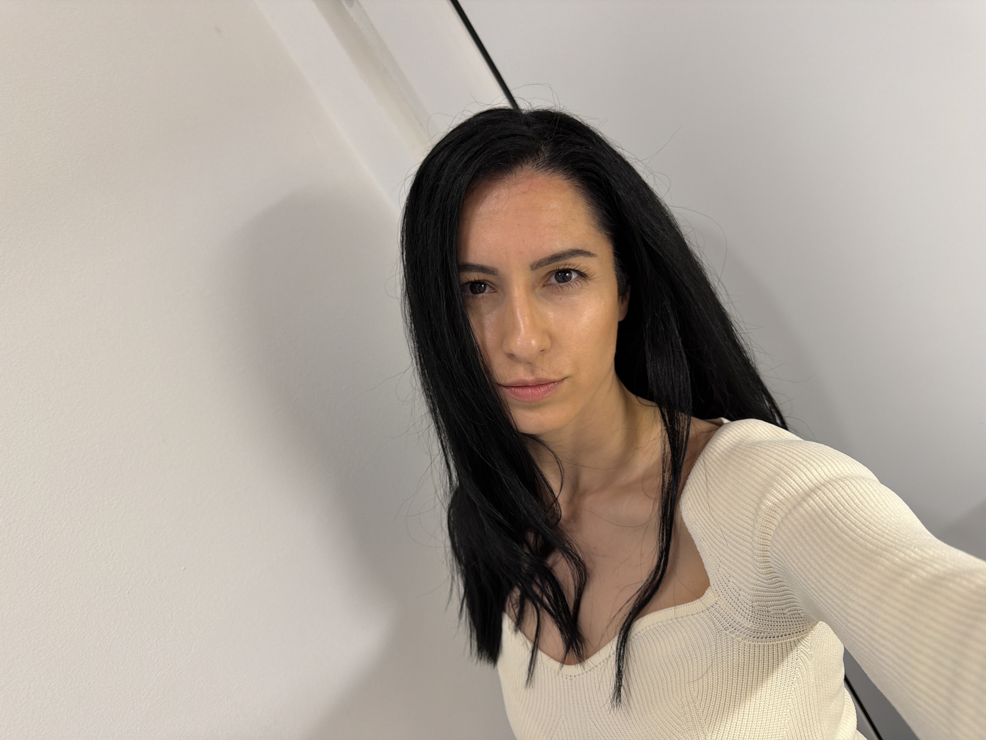 A woman with long, straight black hair is looking directly at the camera. She is wearing a light-colored, long-sleeved shirt with a square neckline. The background consists of a white wall that appears to be a corner, and a black line is visible near the top of the frame. There is no information provided that indicates this image was taken in Pleven, Bulgaria.