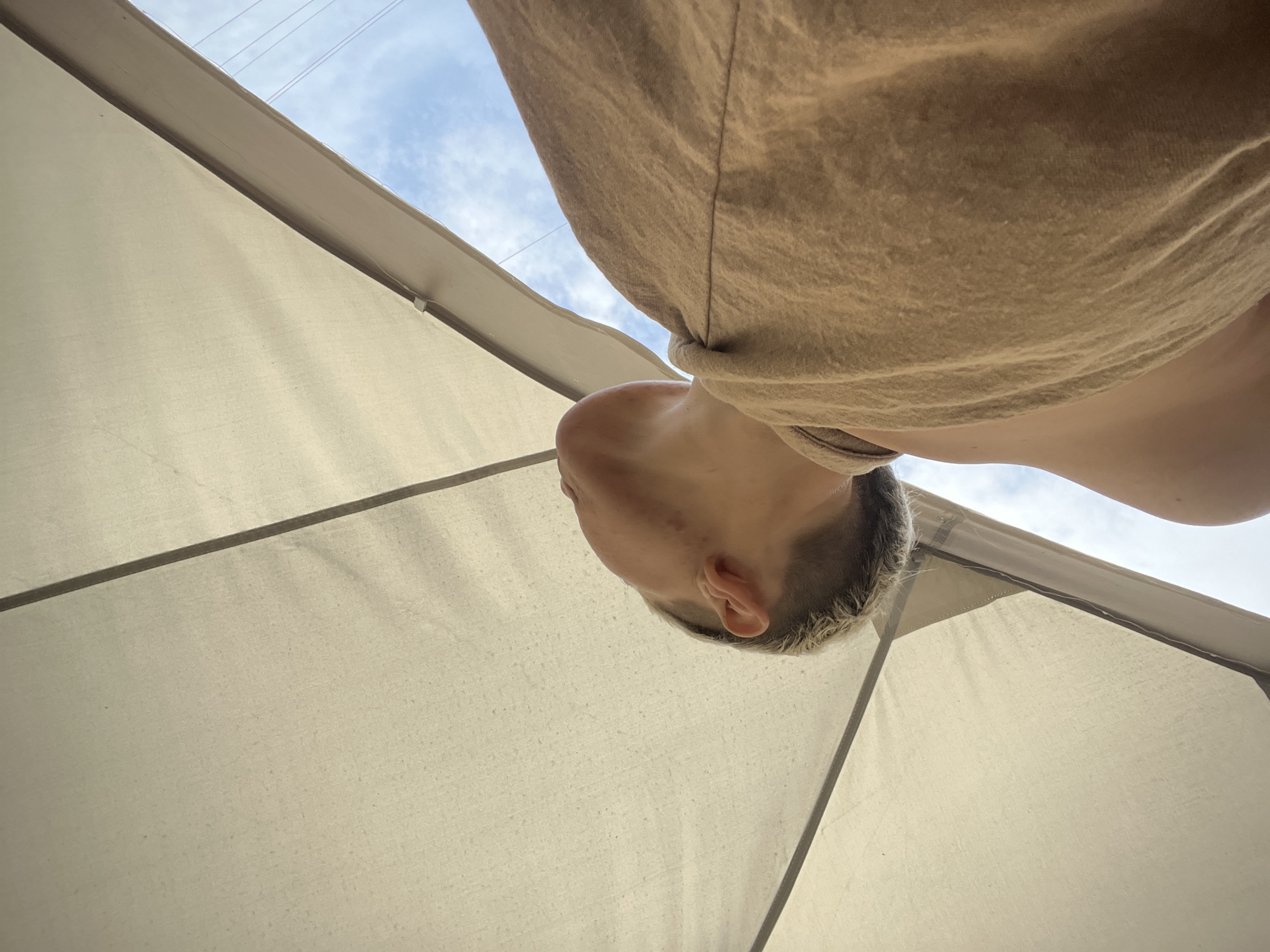 The image shows a low-angle perspective looking upwards. A person, likely a young adult, is visible with their head and upper body taking up a significant portion of the frame. The person has short, light-colored hair, fair skin, and is wearing a light brown t-shirt. The background consists of a white canvas or awning, possibly part of an outdoor structure, with support beams radiating outwards. Above the awning, a small patch of sky with some clouds is visible. It is impossible to determine if the image was taken in the city of Boguchar, Russia.