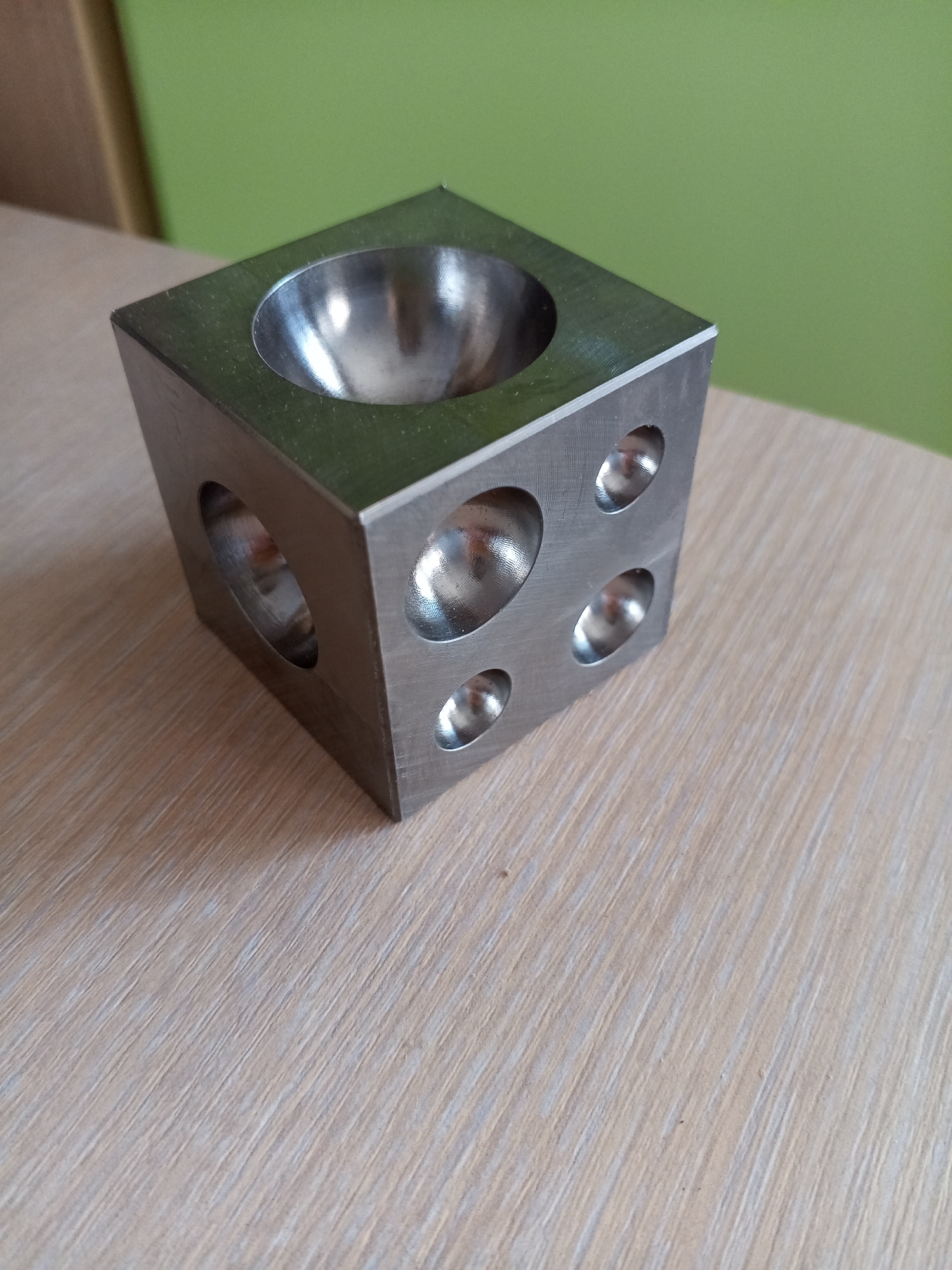 The image shows a metal cube with hemispherical indentations on its faces, placed on a light-colored wooden surface. The cube is positioned at an angle, with one face showing a large concave depression and two adjacent faces each displaying multiple smaller concave depressions of varying sizes. The surface it rests on has a wood grain texture. In the background, a green wall is visible. The scene appears to be indoors, likely on a table. No people or actions are depicted. The object is a forming or dapping block, a tool used in jewelry making and metalworking. The image does not contain information about the city of Vilyuysk, Russia.