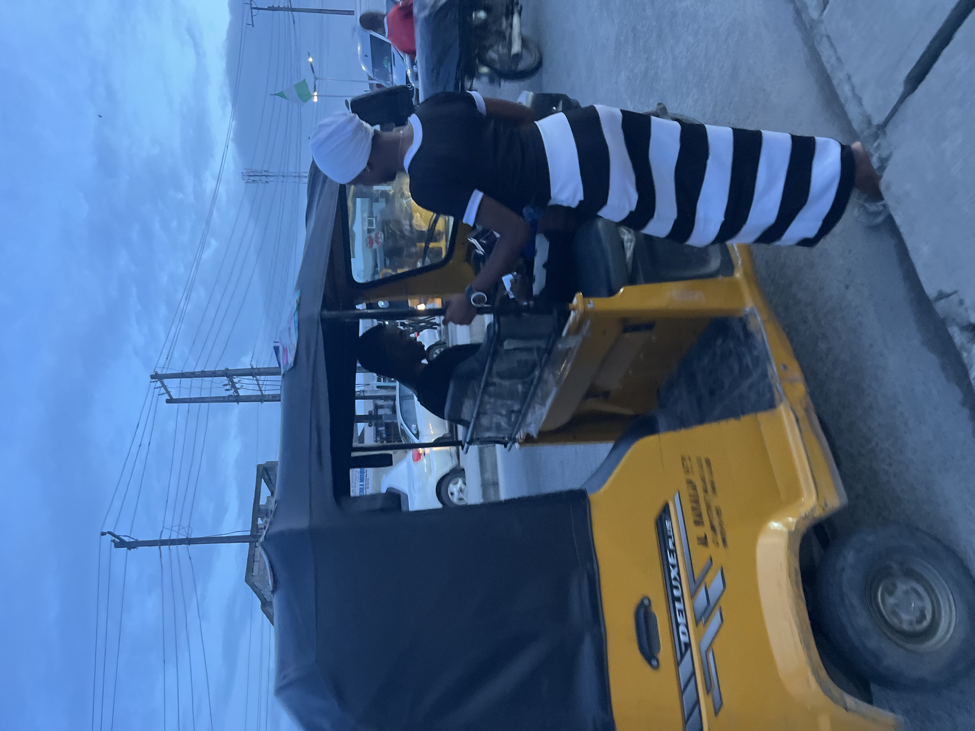 In Ado, Nigeria, at dusk, a yellow motorized rickshaw with a black canopy is parked on a paved surface, likely a street. The rickshaw displays "DELUXE PLUS" and Arabic script on its side. A male driver, wearing a dark shirt, is seated in the driver's position. A woman, wearing a white headscarf and a black and white horizontally striped dress, stands beside the rickshaw, holding its steering wheel. She is barefoot. In the background, a white car is visible, and a Nigerian flag flies from a pole. Overhead, power lines crisscross the sky, which is a pale blue with clouds. A motorcycle with a rider in a red shirt is seen to the right, along with other vehicles and buildings, suggesting an urban environment.
