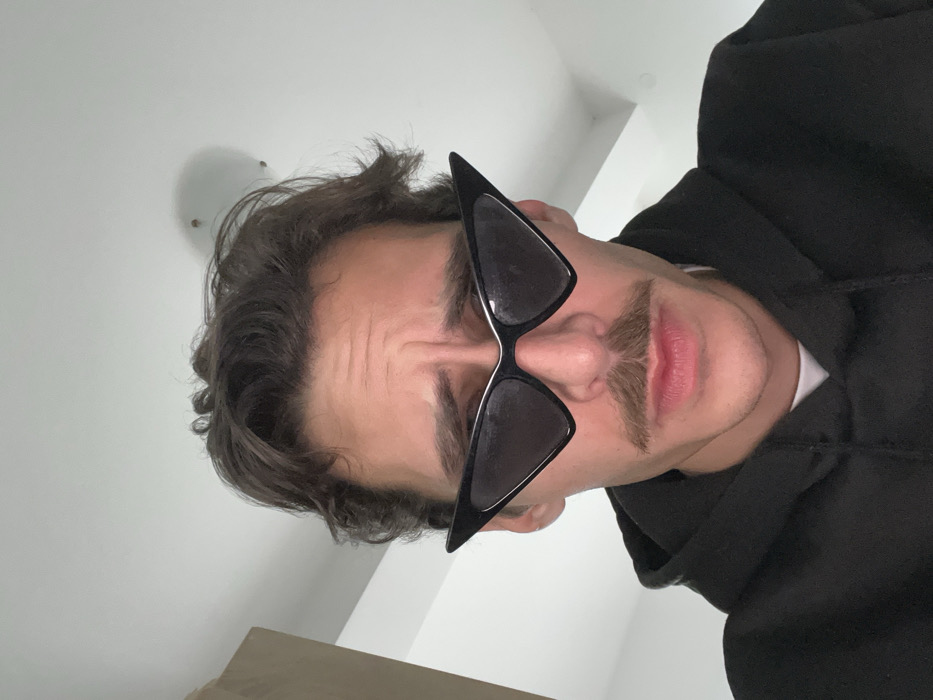 A close-up image depicts a male subject from the chest up, positioned centrally against a light-colored indoor background. The subject, appearing to be an adult male, has medium-length wavy brown hair and a prominent mustache. He is wearing black, sharply angular cat-eye sunglasses with thin silver-colored frames. His eyebrows are furrowed, and his mouth is closed, contributing to a concentrated or slightly serious expression. He wears a black hooded sweatshirt over a white crew-neck undershirt, visible at the collar. The background features a plain, light-colored ceiling with a blurred, circular light fixture partially visible in the upper right quadrant. A light-colored vertical wooden structure is partially visible along the left edge of the frame. The image does not contain any visual information to confirm a specific location in Rafailovići, Montenegro.