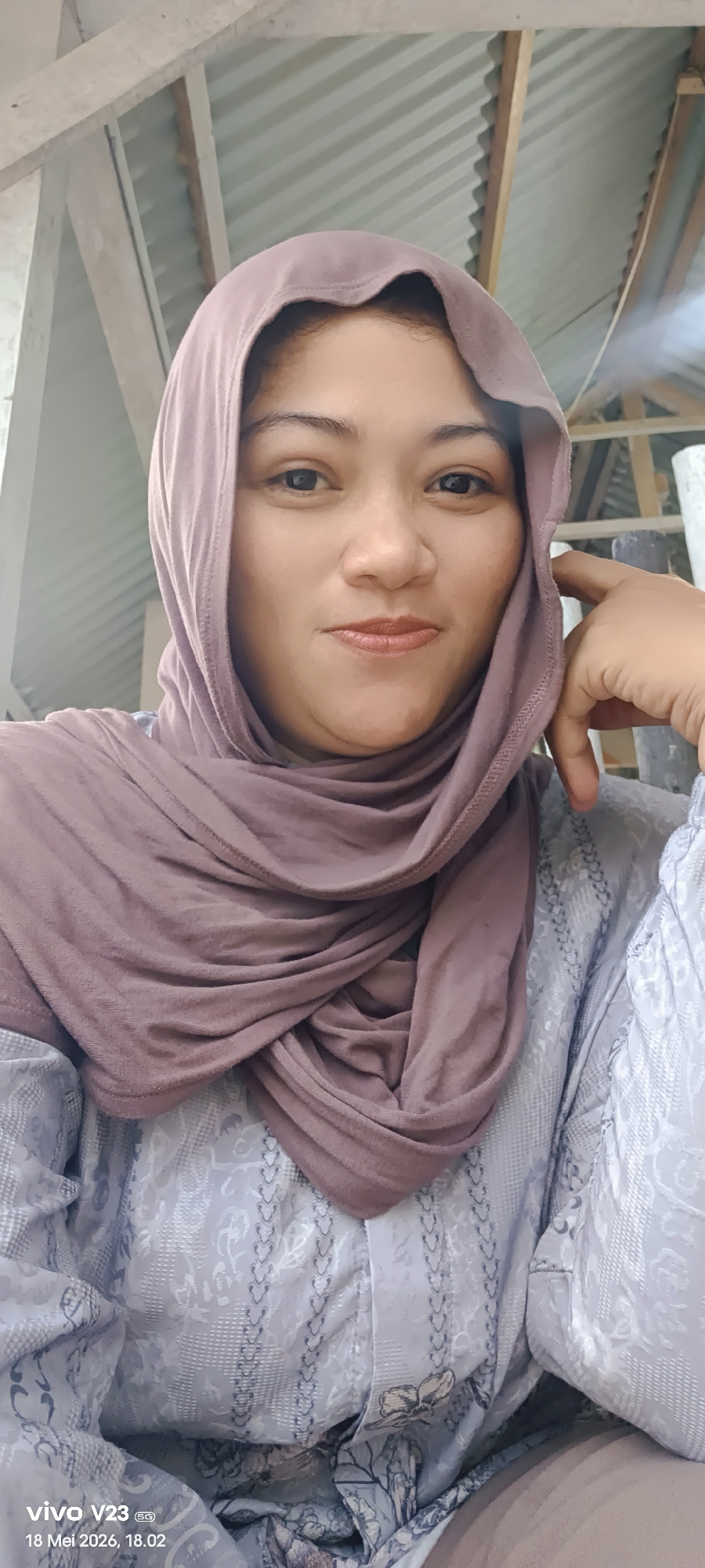 A woman wearing a light purple hijab and patterned blouse is photographed indoors. She rests her head on her right hand, with her fingers slightly touching her face. The background includes wooden structures and corrugated metal, suggesting a building or shelter. The photo was apparently taken using a Vivo V23 smartphone on May 18, 2026, at 18:02. There is no location information provided within Lhokseumawe, Indonesia.