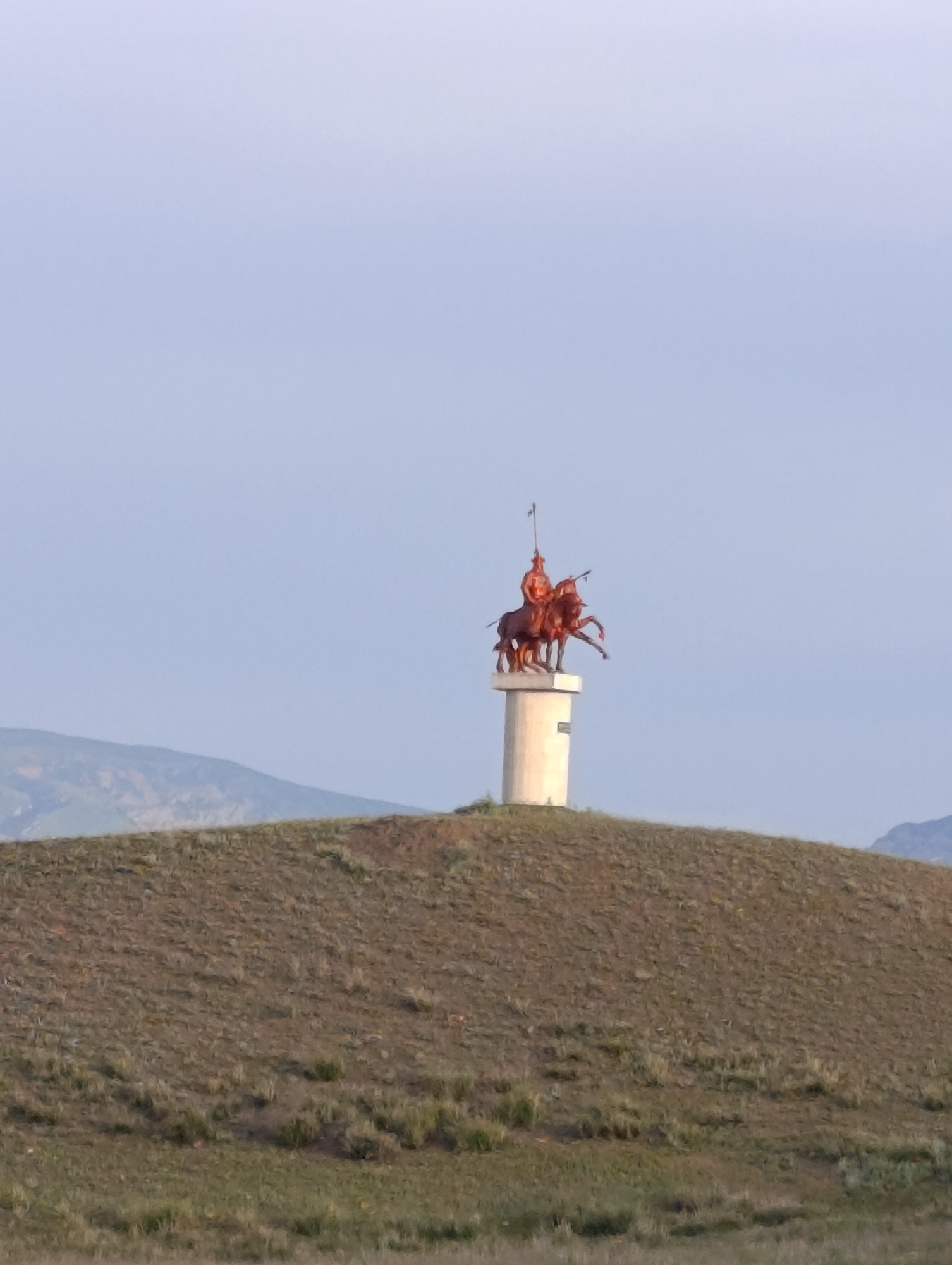The image depicts a statue of two horsemen atop a cylindrical pedestal on a grassy hill. The statue appears to be made of bronze or a similar material with a reddish-brown patina. The horsemen are clad in what appear to be historical or traditional attire, one holding a spear. The pedestal is light-colored and shows some texture, possibly concrete. The hill is covered in dry grass and sparse vegetation, with undulating terrain. In the background, rolling hills and mountains are visible under a clear, pale blue sky. The overall scene suggests an outdoor monument in a natural landscape, likely in a rural or semi-rural setting within the city of Kyzyl-Jyldyz, Kyrgyzstan.