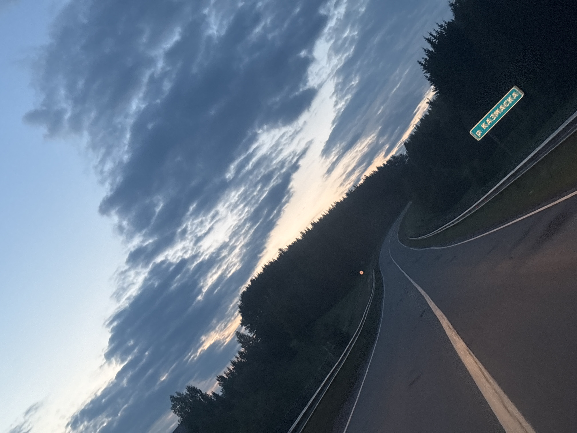 The image depicts a winding asphalt road leading through a forest. A road sign indicates the location as Новая Казмаска (Novaya Kazmaska) in Russia. The sky is filled with streaks of dark clouds with a hint of sunset.