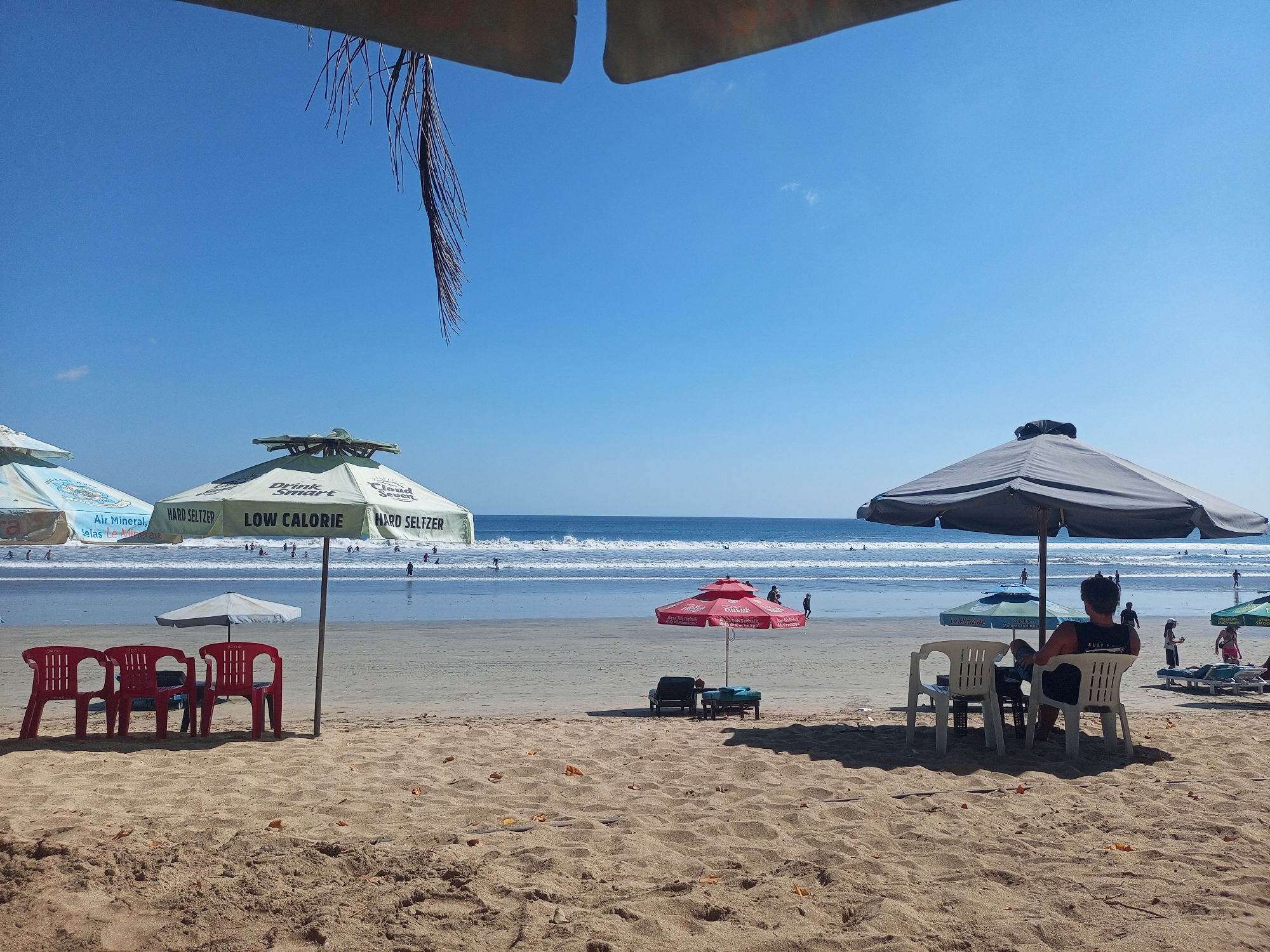 This image depicts a sunny day at Kuta Beach, Indonesia, with a clear blue sky above and gentle waves lapping at the shore. In the foreground, the sandy beach is dotted with scattered dry leaves. Several beach chairs and umbrellas are set up, some occupied and some empty.
On the left side of the image, three bright red plastic chairs are arranged near a large, light-colored umbrella with green accents that reads "HARD SELTZER LOW CALORIE". Further to the left, another umbrella with text visible, including "Air Mineral" and "Le Mineral", is partially in frame.
In the mid-ground, a small red umbrella shades two lounge chairs and a small table. Beyond this, the ocean stretches to the horizon, with several people visible swimming and surfing in the water.
On the right side, a large, dark grey umbrella shades two white plastic chairs where a person is seated, facing the ocean. To the right of this, other beach chairs and umbrellas are visible, and a few people are walking or relaxing on the sand. The overall scene conveys a relaxed beach atmosphere.