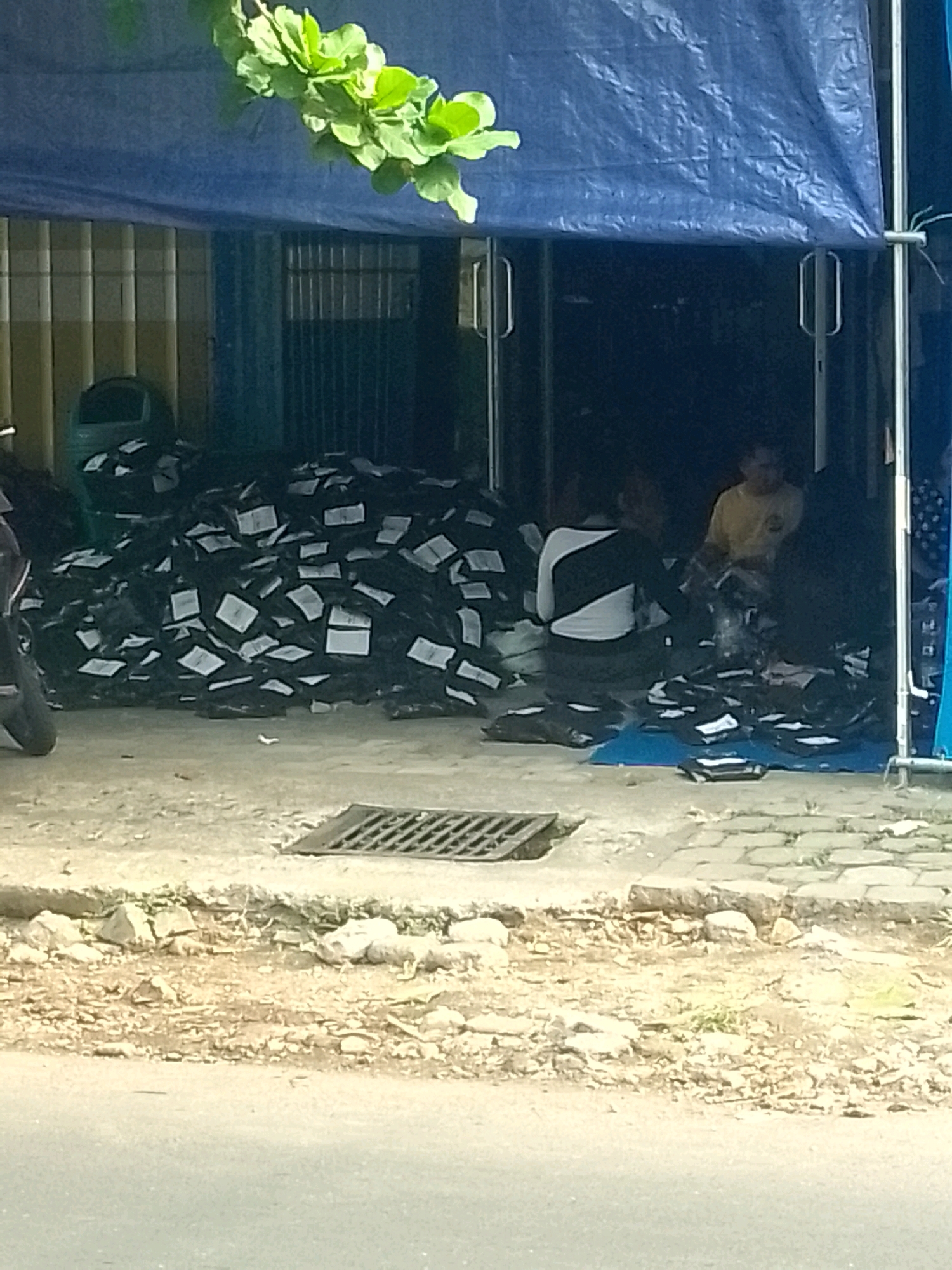 The image depicts an outdoor scene in Gebang, Indonesia, characterized by a large pile of black packages with white labels, suggesting a distribution or shipping point. The packages are spread across the ground and appear to be outside a storefront covered by a blue tarp. A person wearing a yellow shirt is visible inside the storefront, interacting with what appear to be more packages. Another person, wearing a black and white striped shirt, is also present near the packages. A green trash bin is located to the left of the main pile of packages. The ground in front of the storefront is paved with a grate visible in the foreground. Green foliage from a tree overhangs the blue tarp. The overall environment appears to be a commercial area, possibly a small business or warehouse.