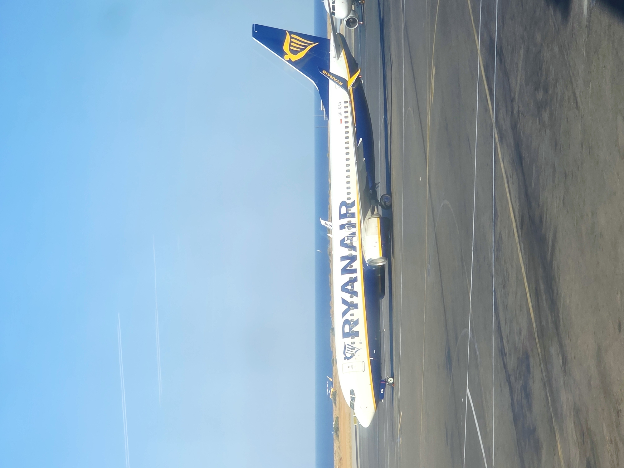 A Ryanair Boeing 737 aircraft is parked on the tarmac at Kos International Airport, also known as Hippocrates Airport, located in the municipal unit of Nea Alikarnassos on the island of Kos, Greece. The plane is white with blue and yellow accents, featuring the Ryanair logo on its tail and "RYANAIR" written in large blue letters along its fuselage. The airport tarmac is grey, marked with white and yellow lines. In the background, the blue sea meets the horizon under a clear blue sky. There are two faint white contrails from other aircraft visible in the distance. The image captures a sunny day, with the aircraft positioned to the right of the frame, slightly angled towards the viewer.