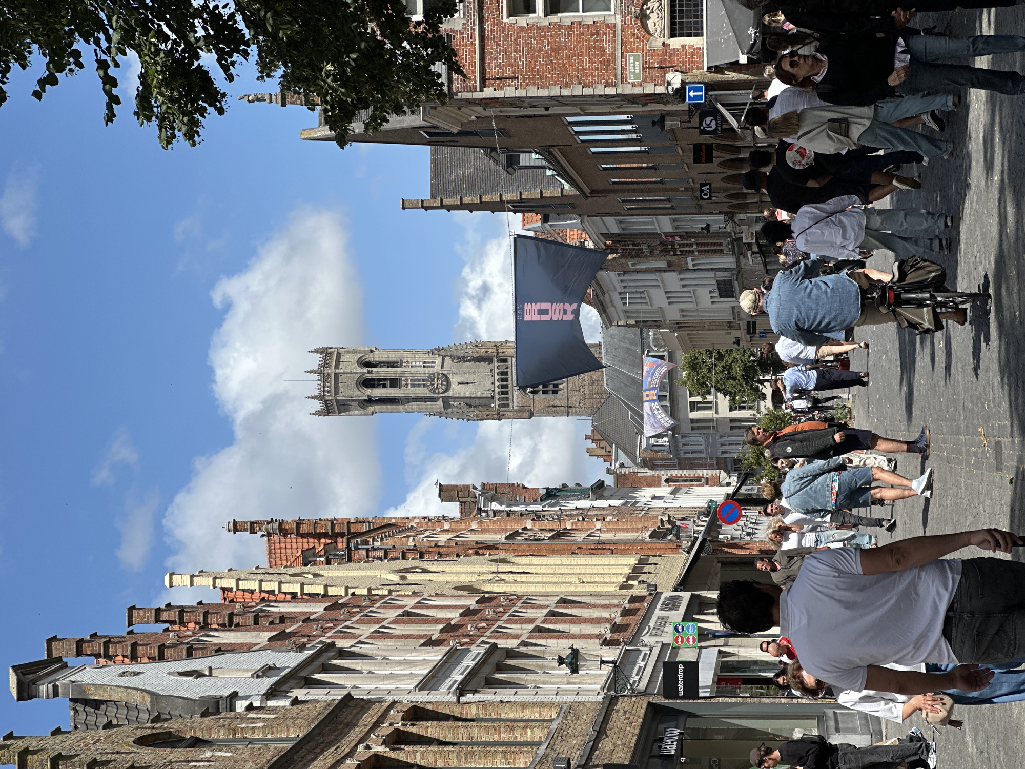 The image depicts a bustling street scene in the city of Bruges, Belgium, featuring the iconic Belfry of Bruges tower in the background. A crowd of people is walking along a cobblestone street lined with tall, narrow, historic buildings with distinct architectural features, including gabled roofs and brick facades. Some of these buildings house shops with visible signage. A large dark banner with pink lettering is suspended across the street between buildings, and a flag with a cityscape graphic is visible further down. The sky is blue with scattered white clouds, and sunlight casts shadows on the street.
In the foreground, a man wearing a white polo shirt and grey jeans is seen from behind, facing away from the camera. To his left, a woman in a white top and blue jeans is partially visible. Further into the crowd, a man in a denim jacket and shorts rides a bicycle, while other individuals are engaged in walking and conversing. The atmosphere suggests a vibrant tourist area.