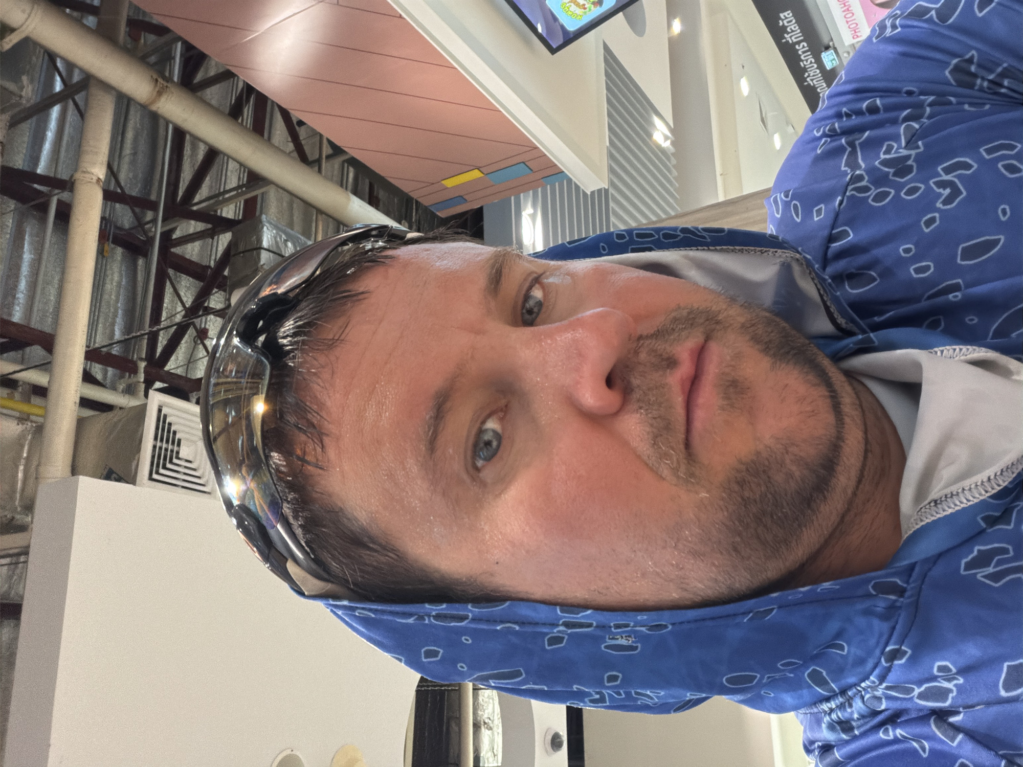 A man with blue eyes, stubble, and a blue hooded jacket with a white pattern is taking a selfie indoors. He is wearing sunglasses on his head. The background shows a ceiling with exposed pipes and ductwork, as well as modern architectural elements that suggest a public indoor space, possibly a shopping mall or transit station. A sign in Thai script is visible in the background, indicating the location is in Ban Bang Rak, Thailand.