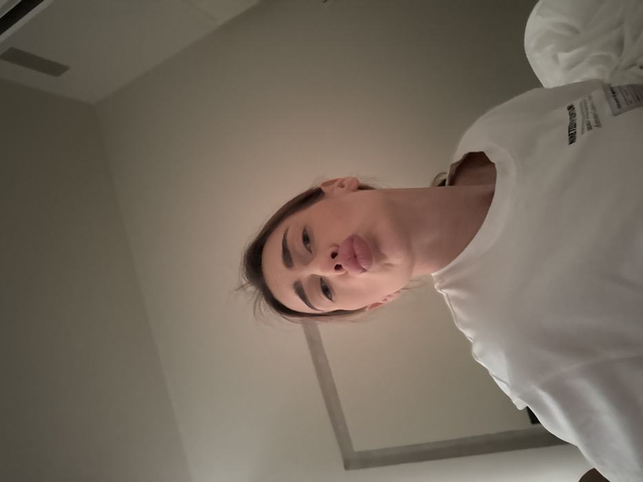 A young adult female is centered in a low-angle selfie, looking directly at the camera with her lips pursed in a "kiss" expression. She has light brown hair, pulled back, and her facial features are clearly visible. She wears a white, short-sleeved t-shirt with dark text and graphics on the lower right front. The environment is an indoor room with light-colored walls and ceiling. A portion of a mirror with a light-colored frame is visible on the left side of the frame, angled towards the left. On the lower right, white crumpled bedding, likely a duvet or sheet, is visible. The upper portion of the image shows a plain, light-colored ceiling, with a subtle corner detail and what appears to be a ventilation grate in the far upper right. The specific location within the city of Venice, Italy, is not discernible from the provided visual information.