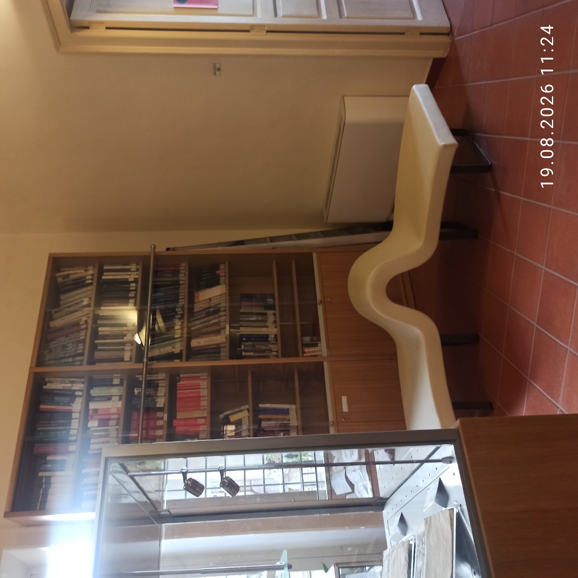 The image shows a room that appears to be a library or study, with shelves filled with books. There is a display case on the left, a unique-looking bench in the foreground, and a door on the right. The floor is tiled in a reddish-brown color. A date and time stamp "19.08.2026 11:24" is visible at the bottom right. The scene is set indoors, with the overall impression of a well-lit and organized space. The location is specified as Ruvo di Puglia, Italy.