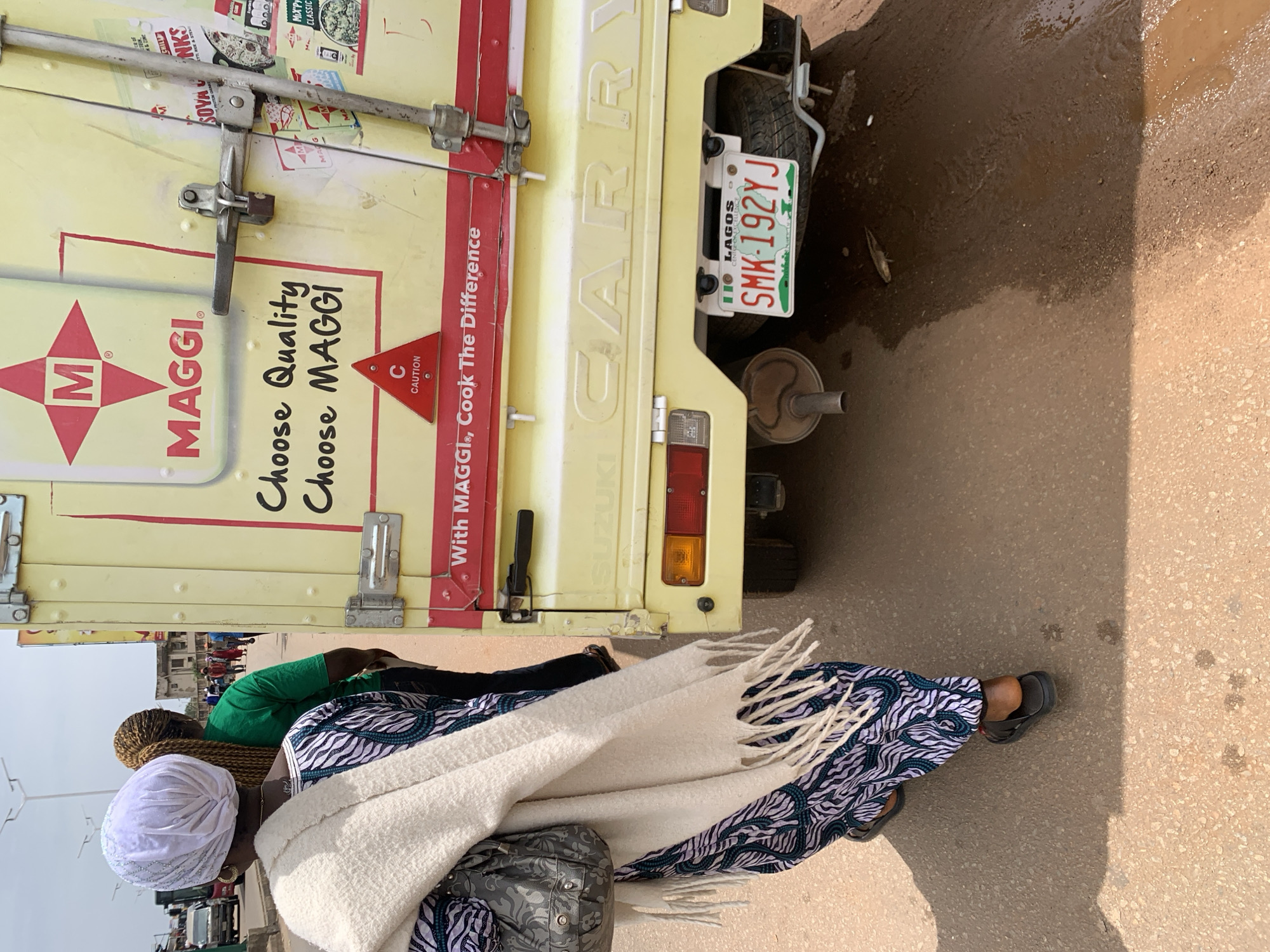 The image depicts a street scene in Jos, Nigeria. A yellow delivery truck, branded with "MAGGI" and the slogan "Choose Quality, Choose MAGGI", is parked on a dusty, paved road. The truck has "SUZUKI CARRY" embossed on its rear. A license plate reading "LAGOS SMK-192YJ" is visible. A woman in a patterned blue and white dress, a white headscarf, and a cream-colored shawl is walking away from the camera, towards the left side of the frame. She is carrying a grey patterned bag on her left shoulder. Behind her, another woman wearing a green shirt with her hair in braided extensions is also walking away. The road surface appears worn and somewhat muddy in places, suggesting recent rain or poor drainage. Other vehicles and buildings are visible in the background, indicating an urban environment. The overall impression is that of a typical street scene in a Nigerian city, with commercial activity and pedestrian traffic.